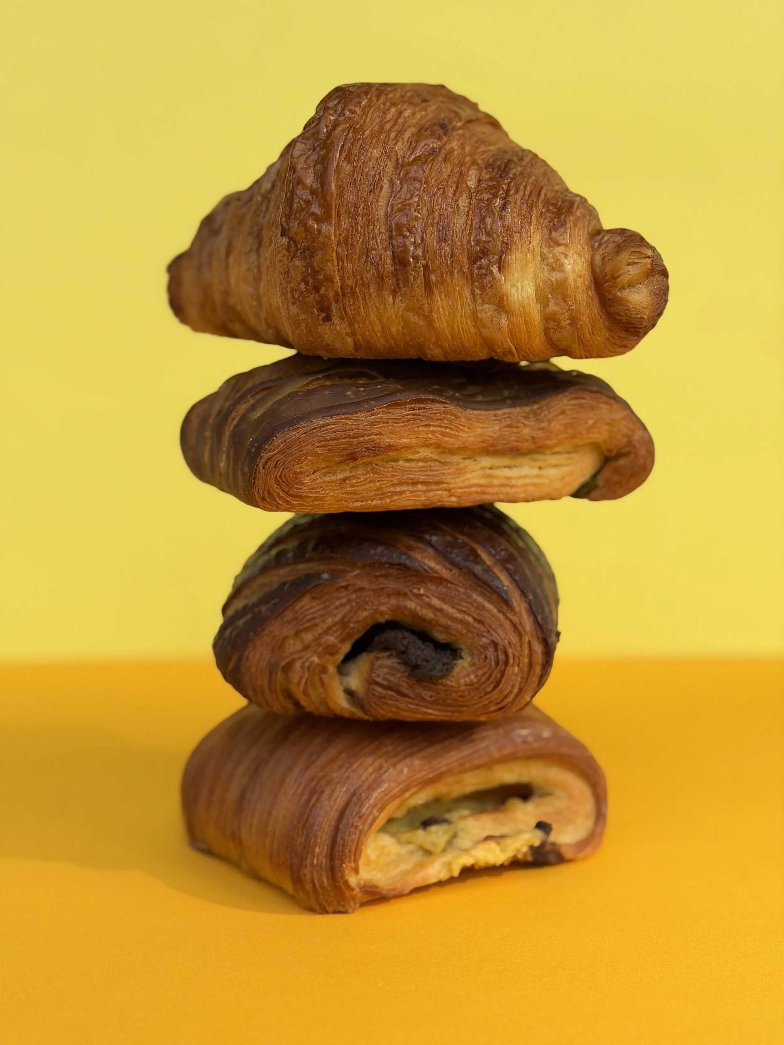 A stack of croissants with the bottom one cut open, showing a filling of scrambled eggs and chocolate chips, against a yellow background.