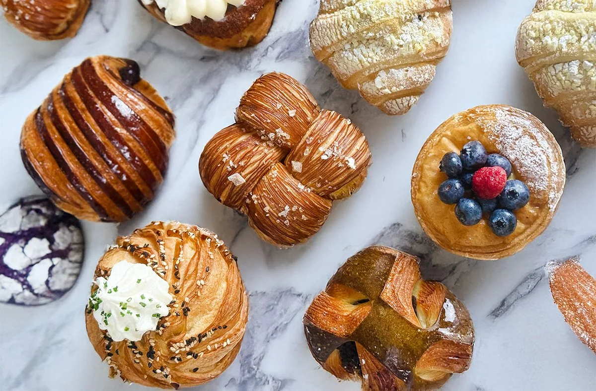 Assorted pastries and small desserts on a white marble surface, including croissants, a fruit tart topped with blueberries and a raspberry, a pastry with whipped cream, and a chocolate-drizzled pastry.