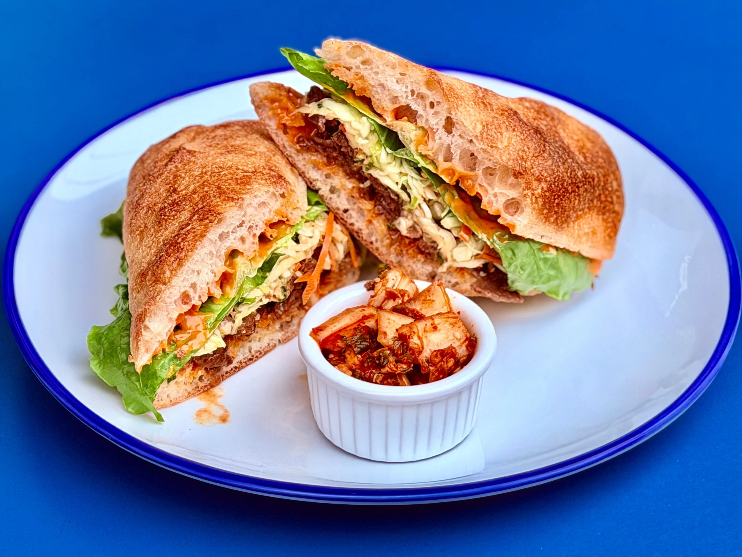 Close-up of a half-eaten burrito with lettuce, shredded carrots, and meat, served with a side of kimchi in a white ramekin on a white plate with a blue rim, on a blue surface.