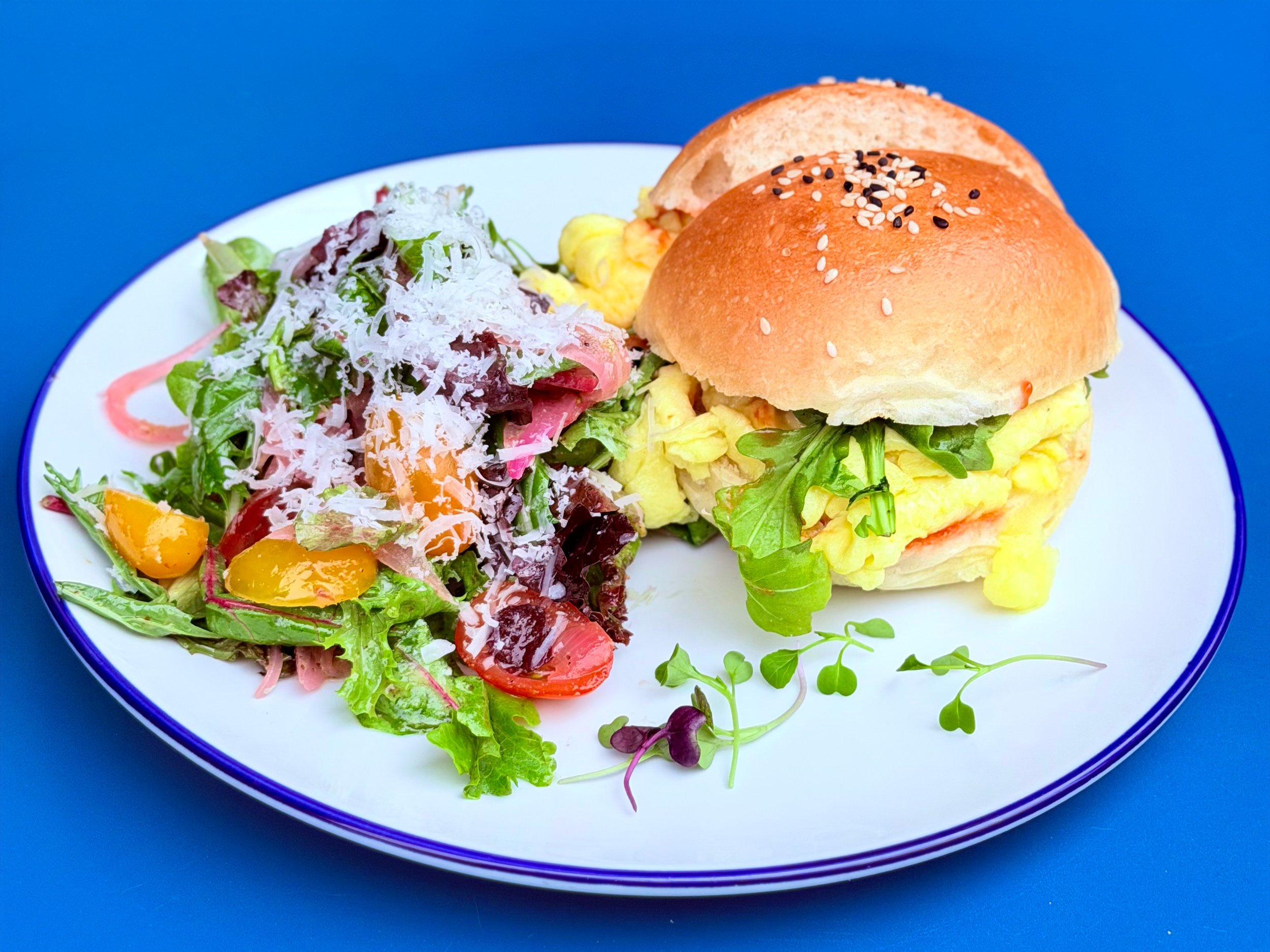 A white plate with a blue rim features a sandwich with scrambled eggs, lettuce, and a roll bun, accompanied by a side salad with mixed greens, cherry tomatoes, grated cheese, and pink dressing, set against a blue background.