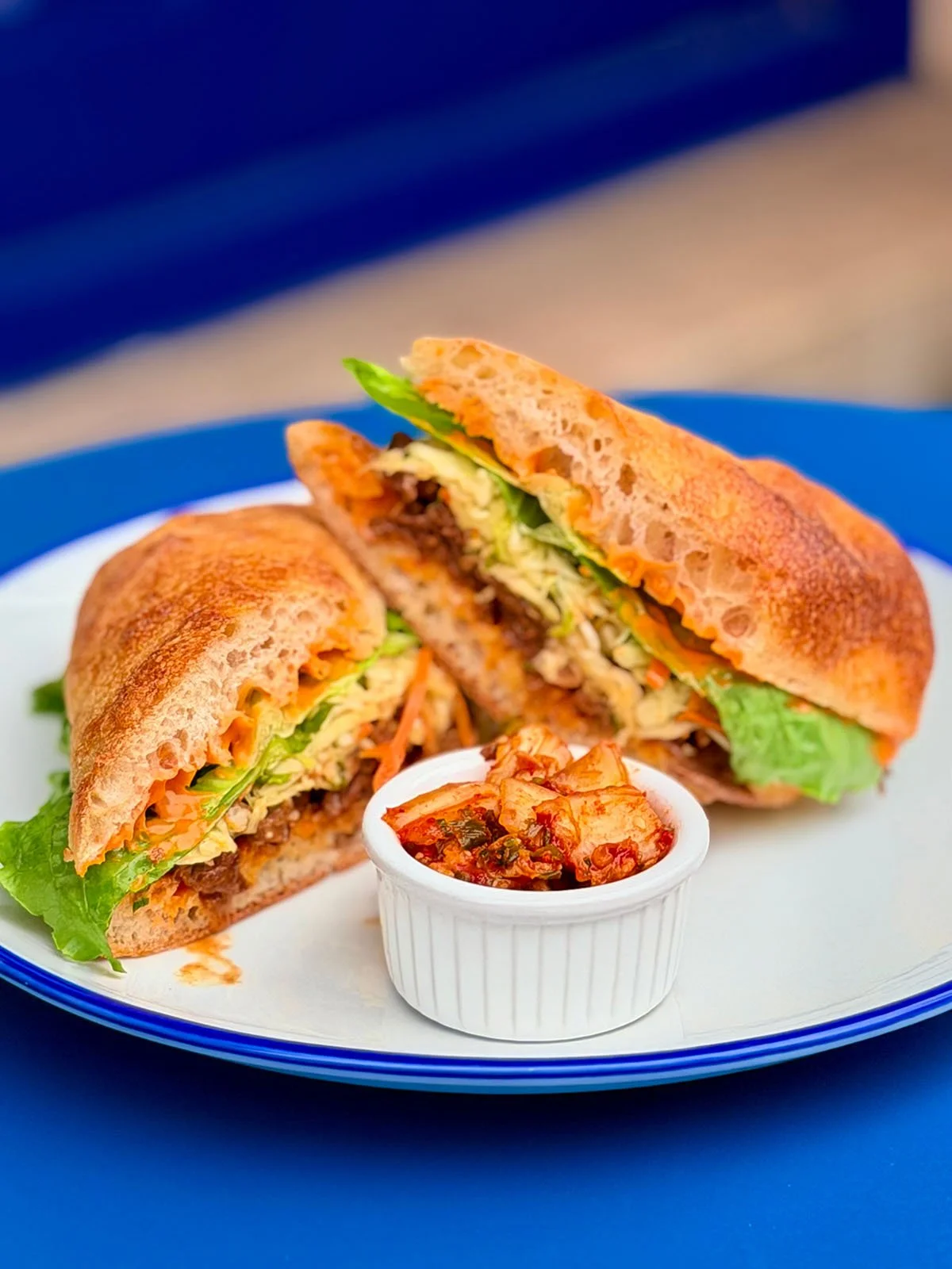 A sandwich cut in half with lettuce, meat, and vegetables inside, served with a side of kimchi in a white ramekin on a blue-rimmed plate.