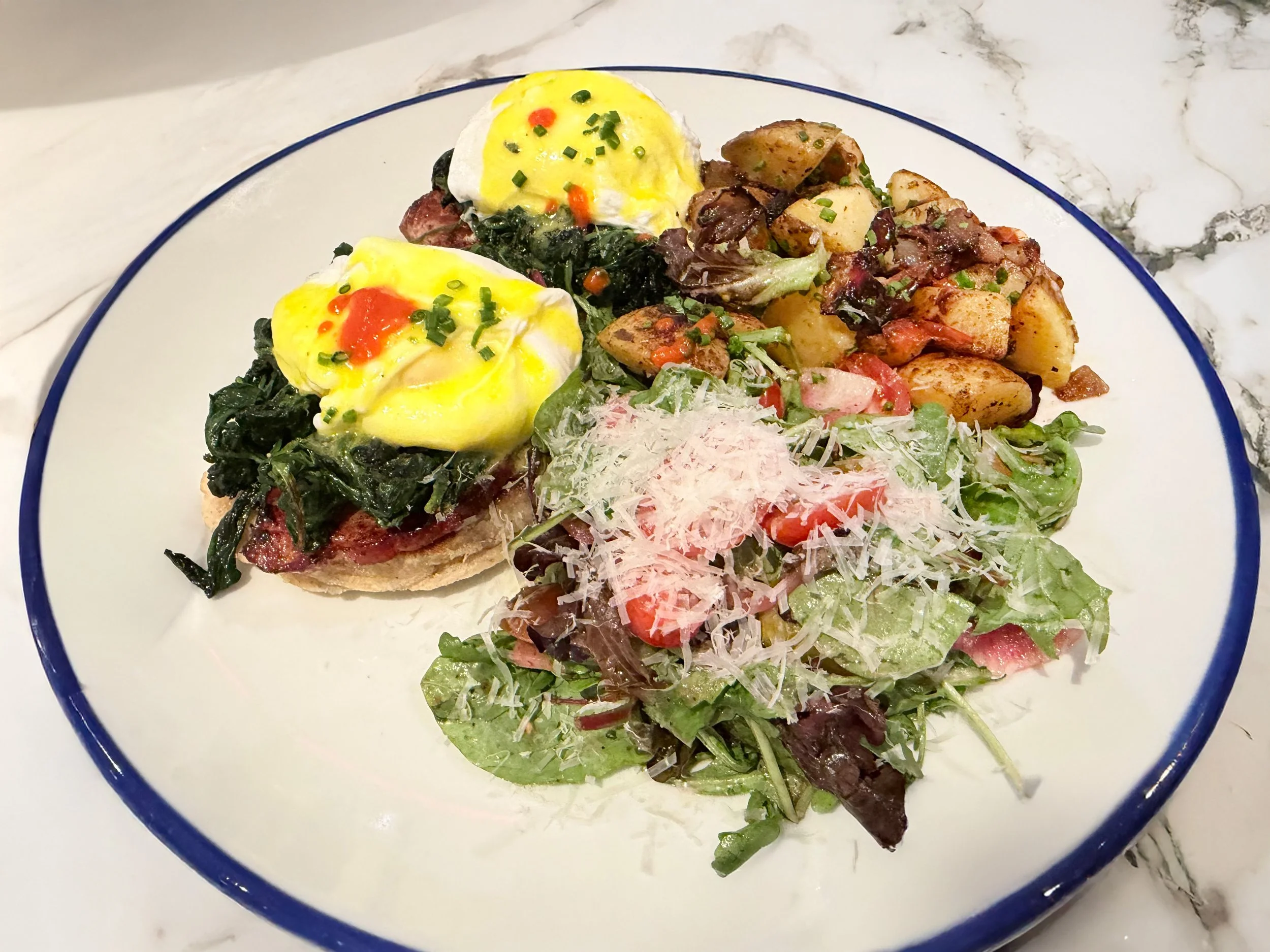 Plate of food with poached eggs on toast, roasted potatoes, mixed greens, and salad with shaved cheese.