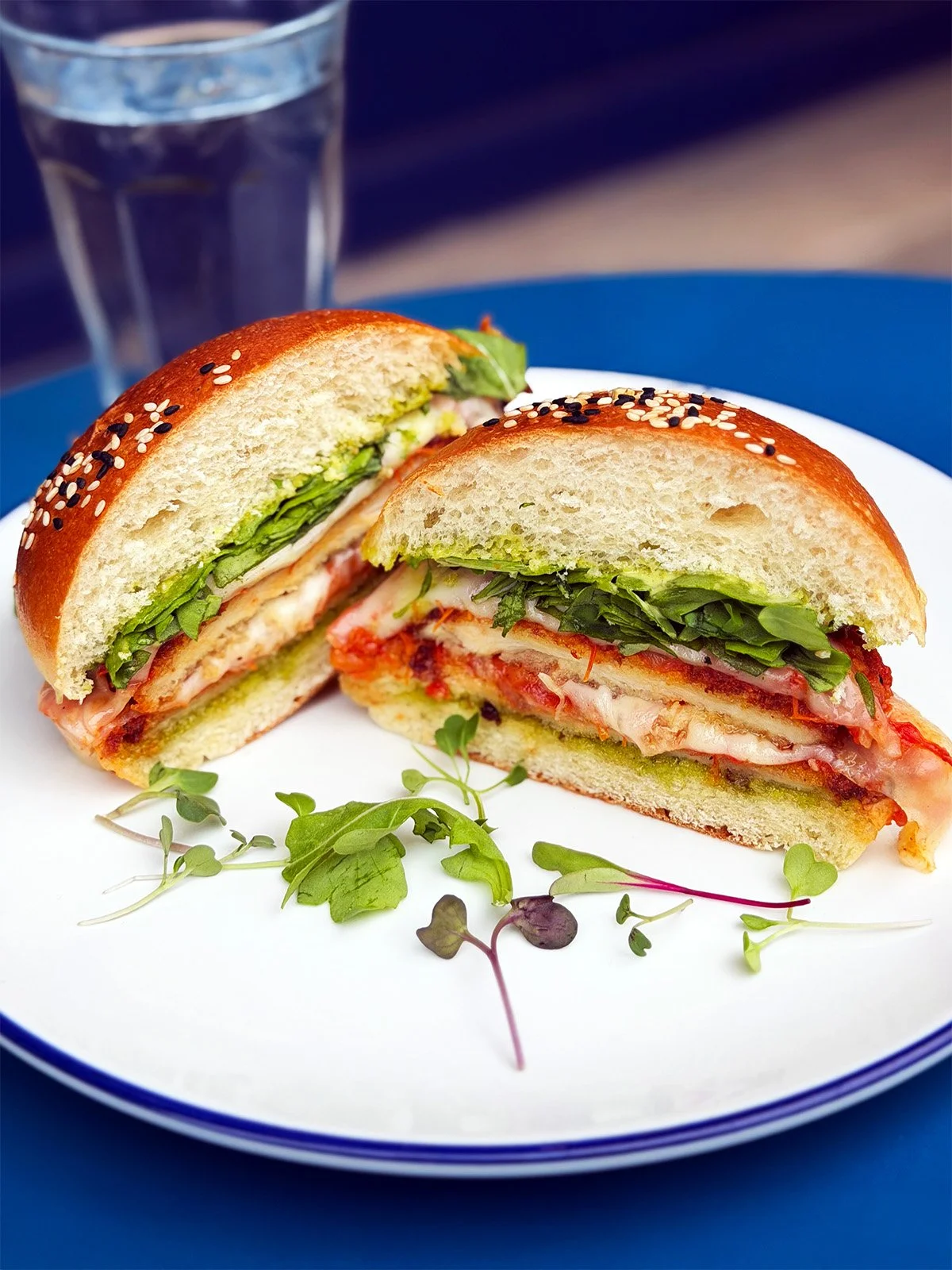 A close-up of a sandwich with lettuce, tomato, cheese, and deli meat in a sesame seed bun, cut in half and placed on a white plate with microgreens and an ice water glass in the background.
