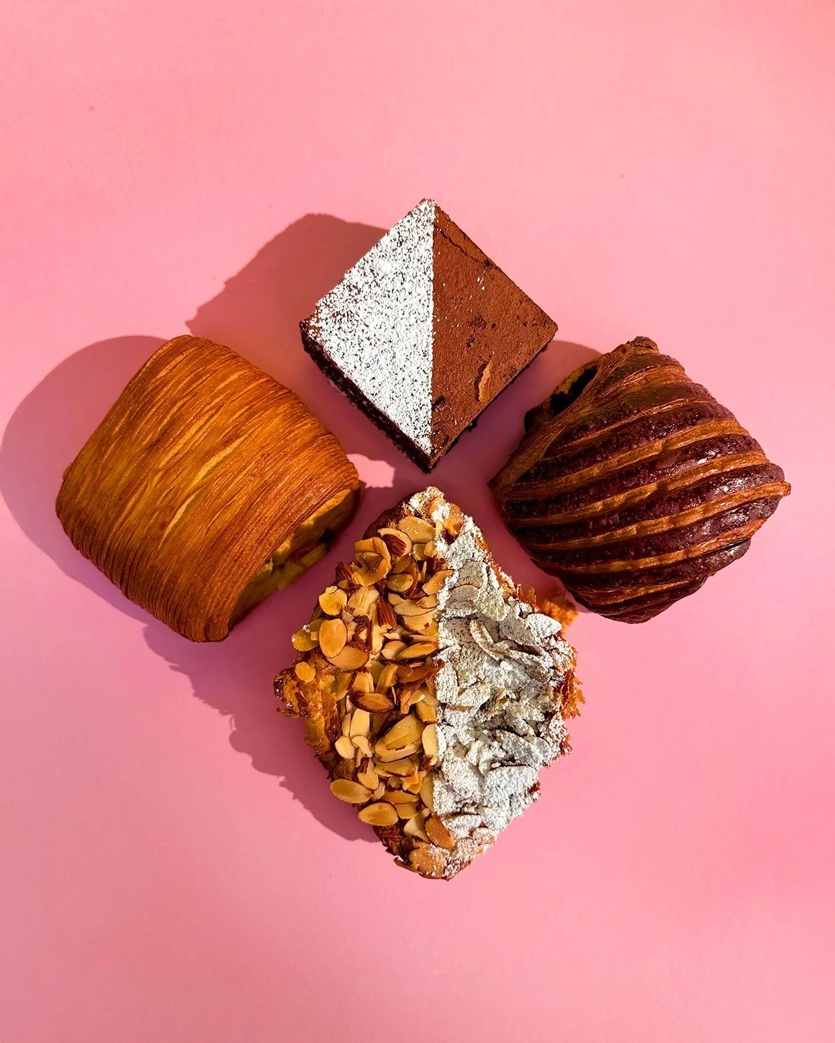 Four assorted pastries arranged on a pink surface, including a square chocolate cake with powdered sugar, a layered puff pastry, a chocolate and caramel swirl pastry, and a sliced pastry topped with powdered sugar and sliced almonds.