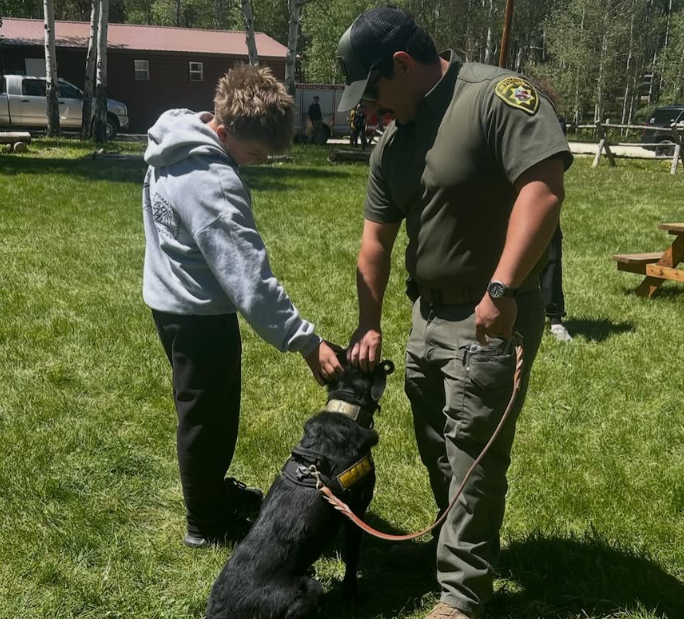 A person in a police uniform interacts with a young boy and a black service dog on a grassy field in a park.