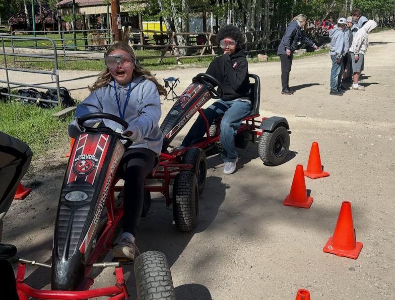 Two children riding in a red go-kart with a black helmet design, one girl in the front wearing glasses and a gray hoodie, another boy sitting behind her, both smiling. Orange traffic cones line the dirt track on a sunny day, with people and trees in the background.