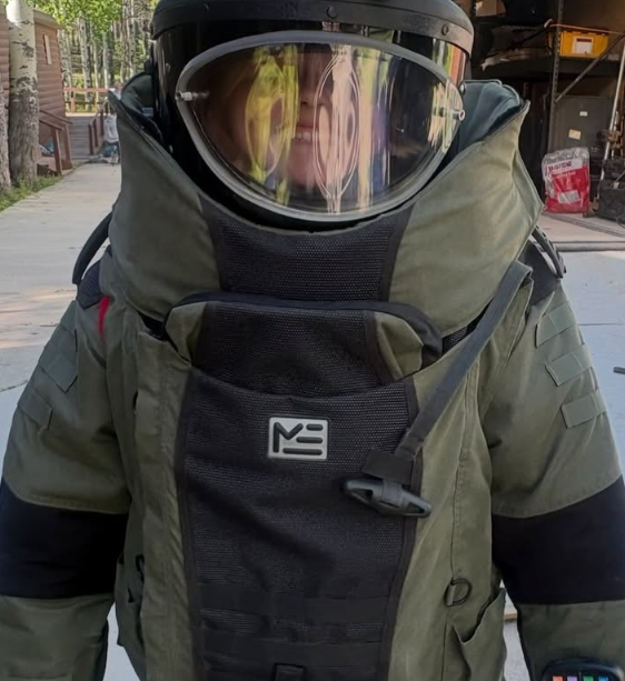Person wearing military-style tactical gear, including a helmet with a reflective visor, a green tactical vest, and black long sleeves.