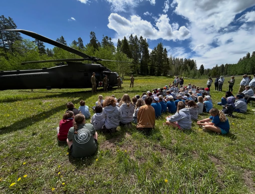 A group of children sitting on grass and listening to a presentation by military personnel near a black helicopter in a green outdoor field with trees and a blue sky.