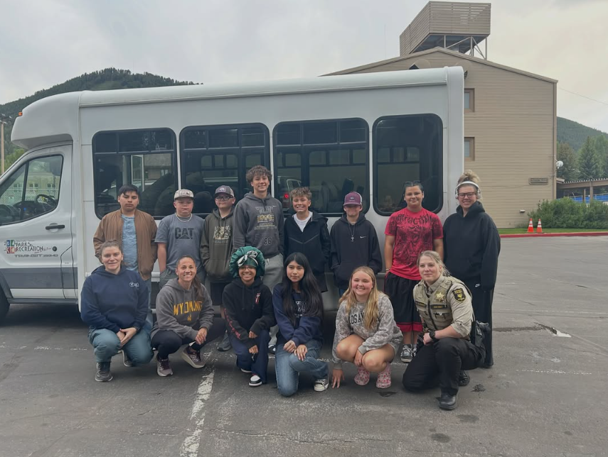 Group of young people, including children and teenagers, posing in front of a white bus outdoors with a building, mountains, and cloudy sky in the background.