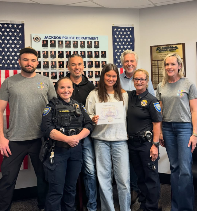 Group of six people, including two police officers and a young girl holding a certificate, posing in front of a police department board and American flags.