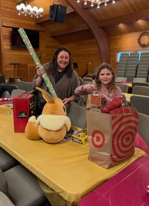 A woman and a young girl standing behind a table filled with gifts in a wooden room, likely for Christmas. The woman is holding a large wrapped gift, and the girl is holding a smaller gift. On the table are a large plush bunny, a red box, a small yellow and black box, and a target shopping bag with gifts inside.