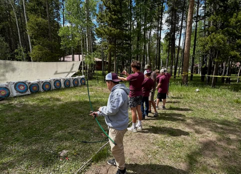 Group of children and teenagers practicing archery outdoors in a wooded area, aiming their bows at targets.