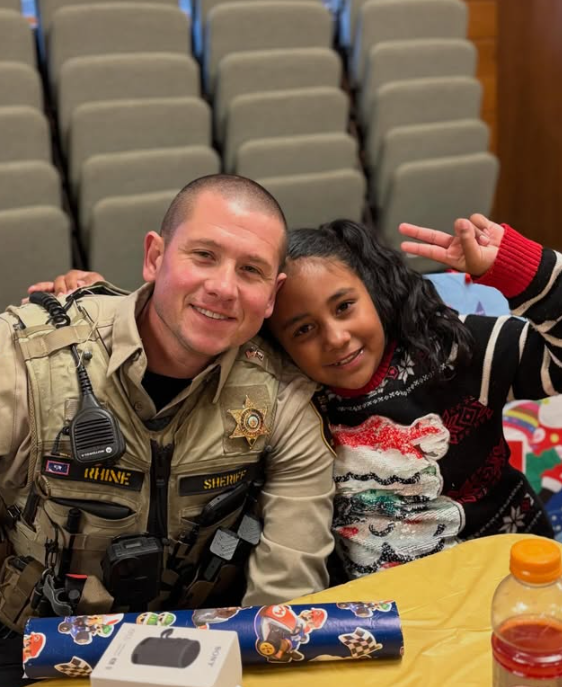 A smiling uniformed sheriff's deputy with a badge and radio, sitting next to a young girl with curly hair, both making peace signs, in a room with rows of empty theater seats behind them.