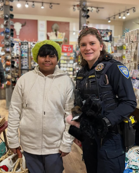 A young boy wearing a bright green knit hat and a beige hoodie standing next to a female police officer in a retail store. The officer is in uniform, smiling, holding a small black puppy, and there are shelves of products in the background.