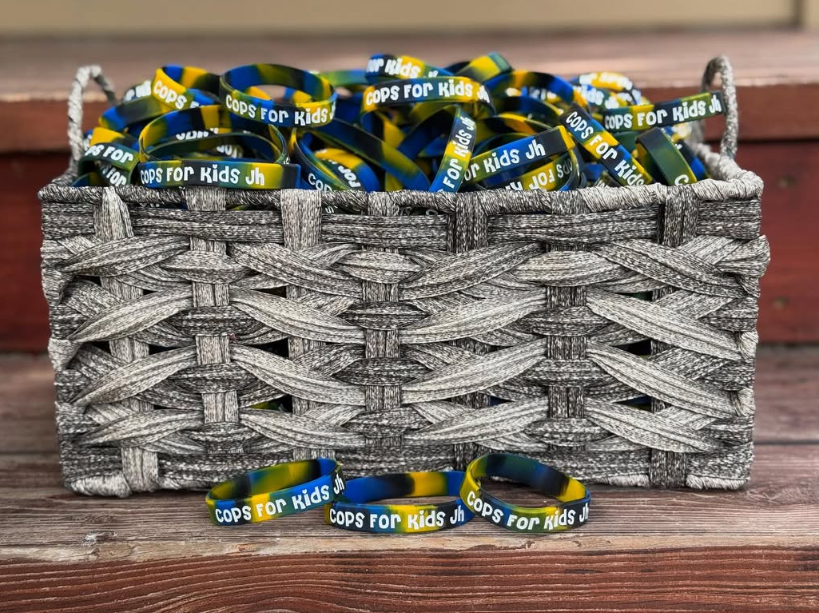 A woven gray basket filled with yellow, blue, and white silicone wristbands that read "Cops for Kids JH," with a few wristbands laid out in front of the basket on a wooden surface.