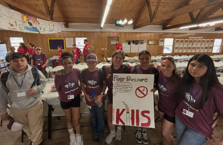 A group of six young people standing together inside a wooden cabin, holding a sign that reads 'Peer Pressure Kills', with some additional drawings and a crossed-out cigarette symbol.