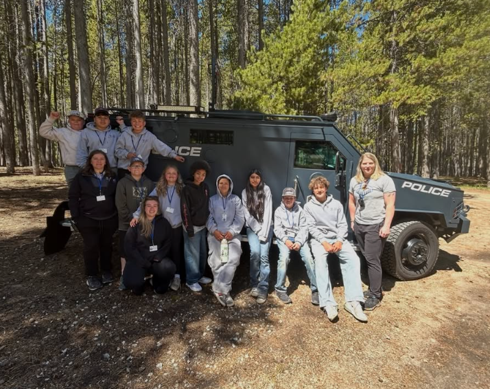 Group of children and a woman standing in front of a police vehicle in a forested area.