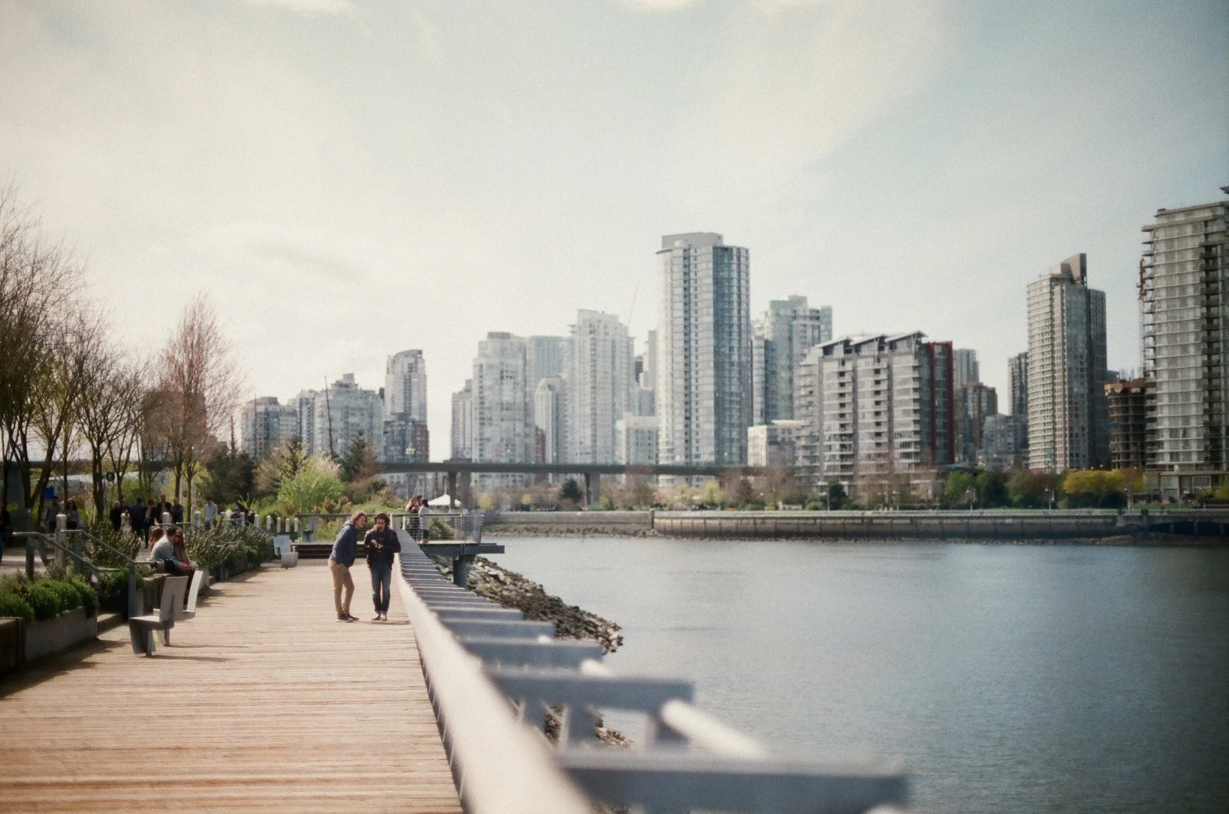 People walking and sitting on benches along a waterfront promenade with a city skyline of tall buildings behind