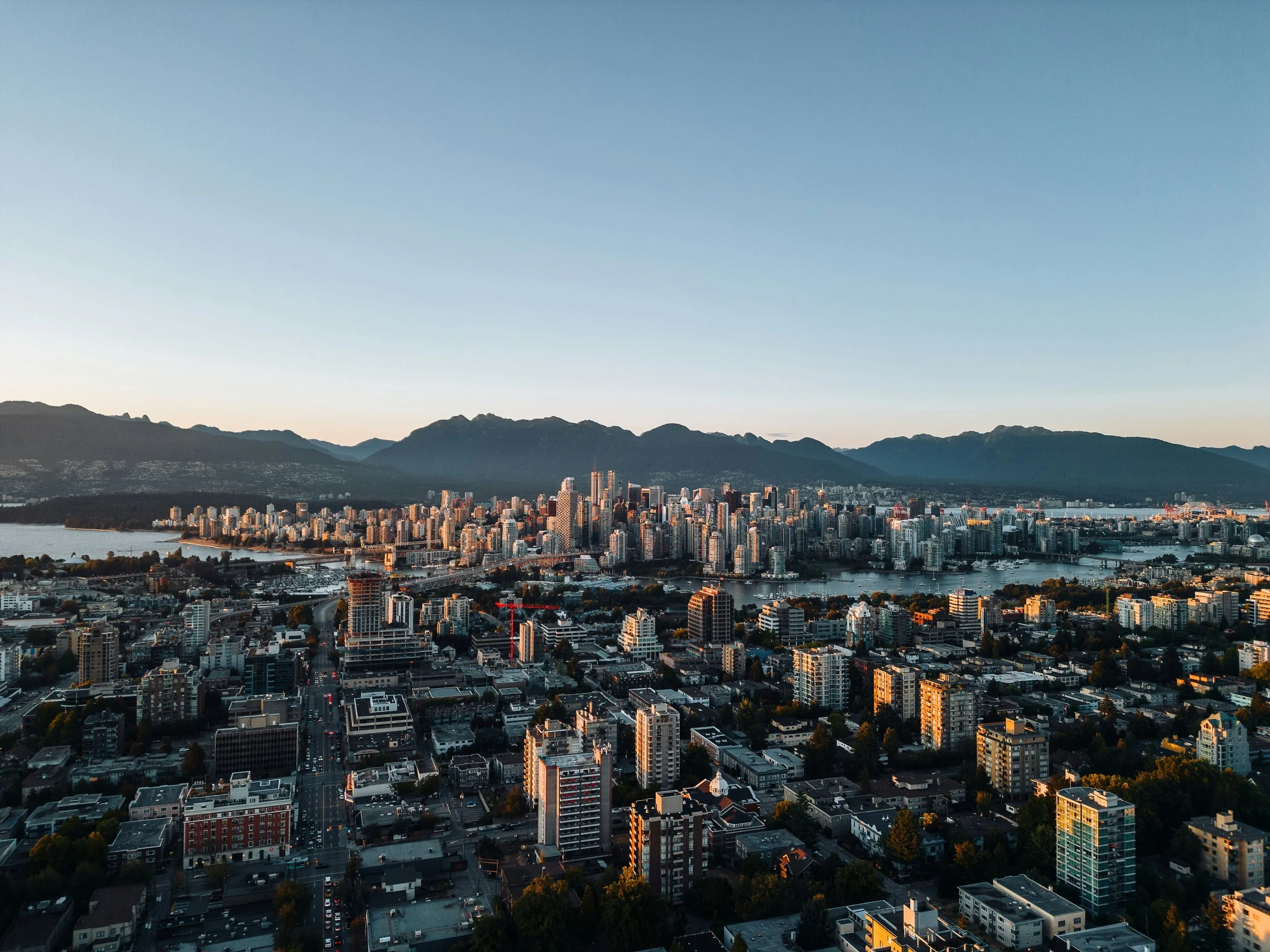 Aerial view of a cityscape with high-rise buildings, water, and mountains in the background at sunset.