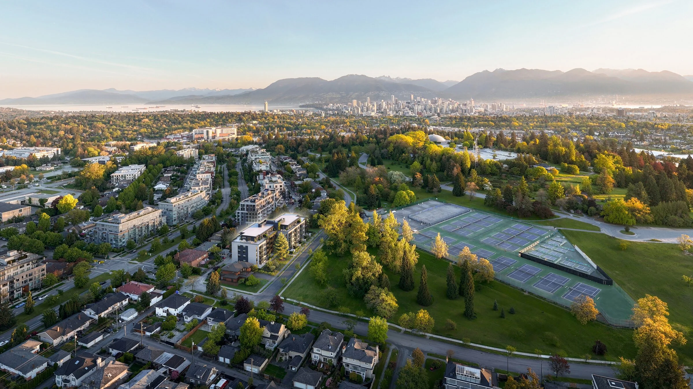 Aerial view of a city landscape with a park, tennis courts, residential houses, and tall buildings in the distance with mountains and a body of water in the background during sunset.