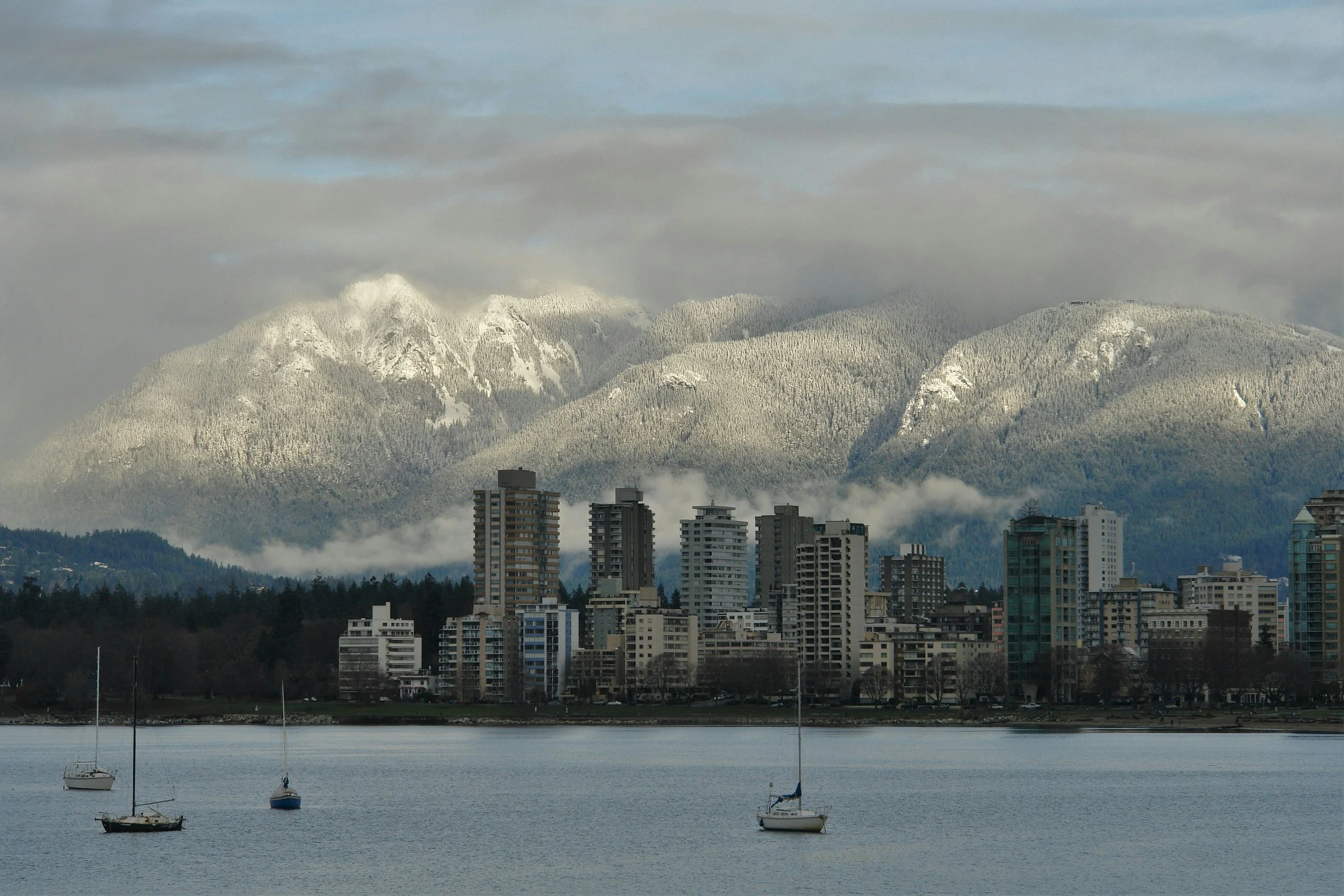 City skyline with high-rise buildings along a waterfront with sailboats, mountains in the background covered in snow, and cloudy sky.