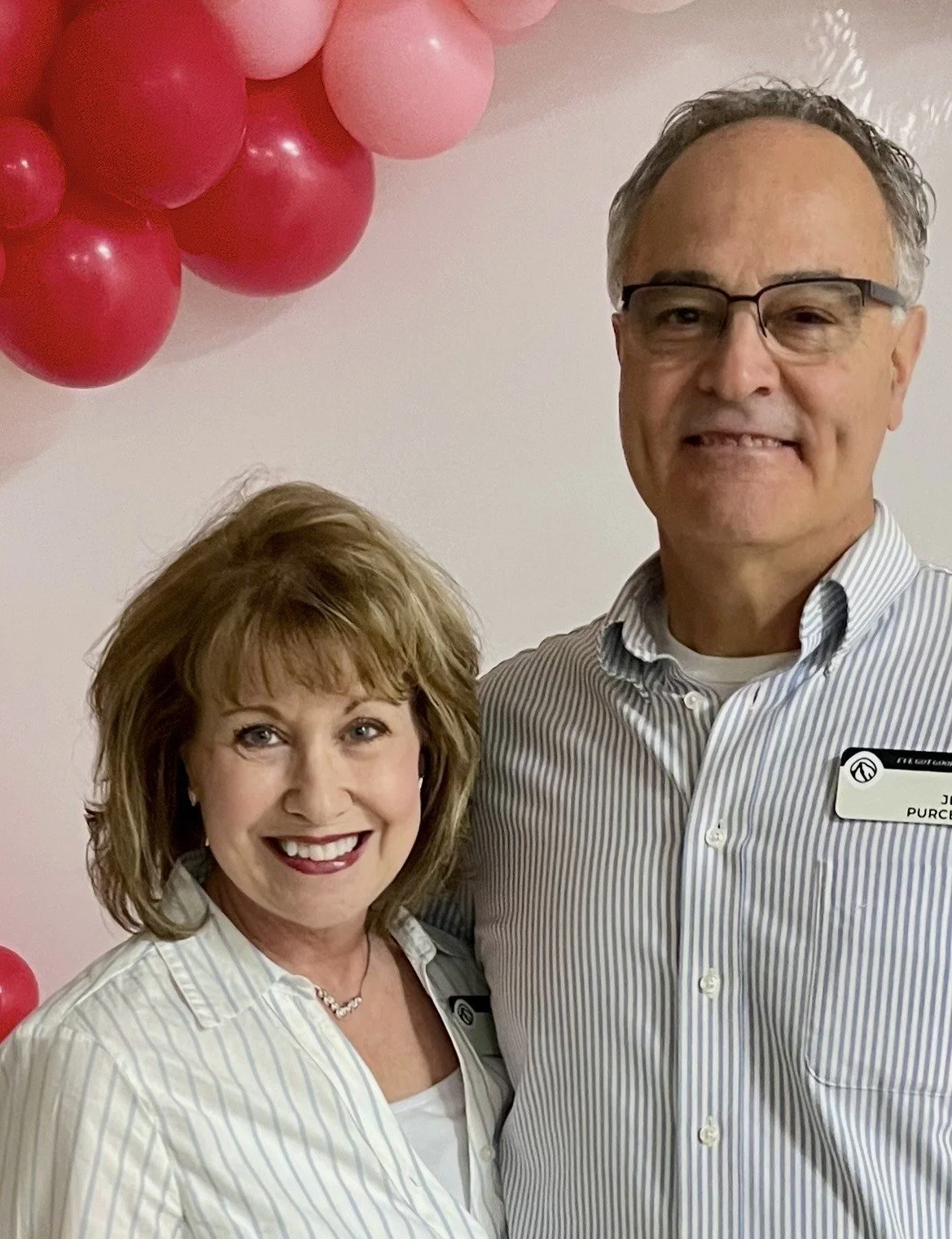 A smiling middle-aged couple standing outdoors with trees and cars in the background.