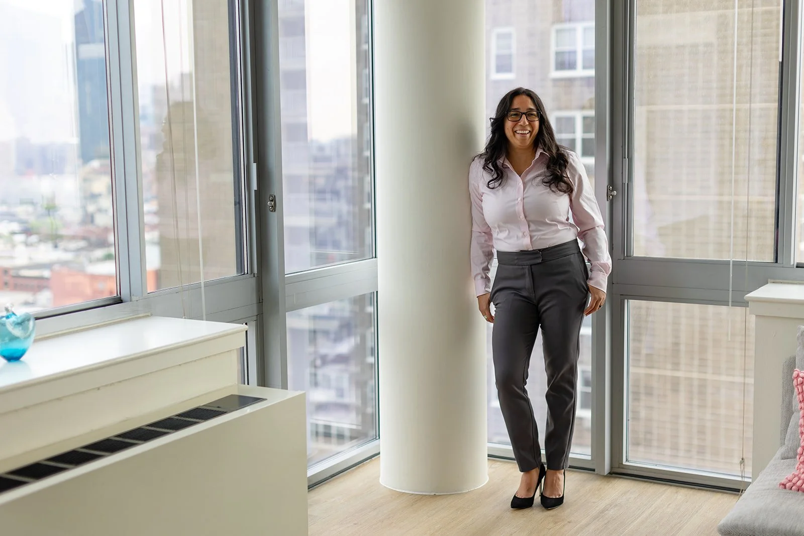 A woman with glasses, wearing a pink shirt and dark gray pants, stands near a large window in a high-rise apartment or office, smiling.