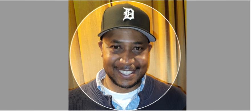 A man smiling, wearing a black baseball cap with a white letter D, and a dark sweater over a light blue shirt, standing in front of a yellow curtain.