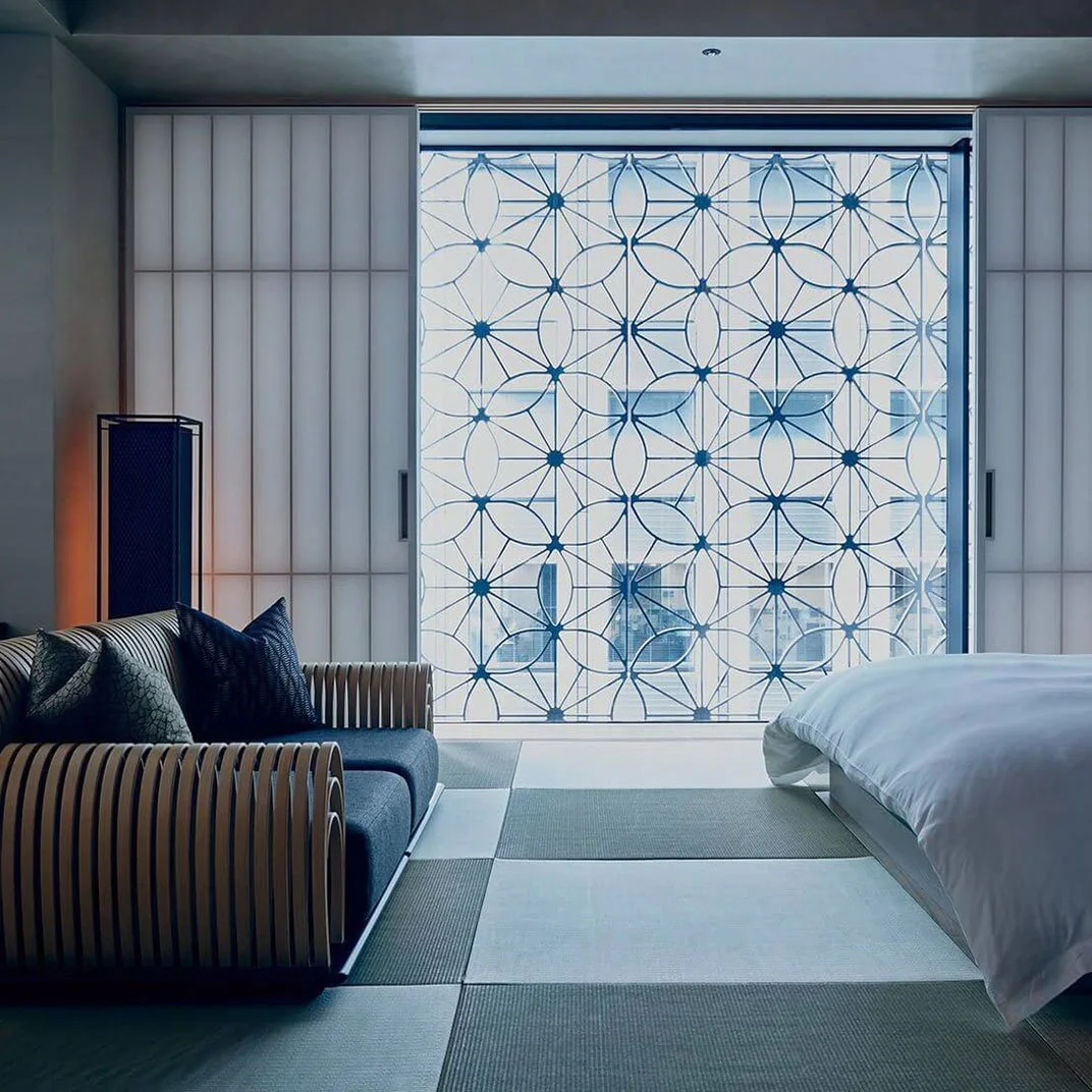 Modern hotel room with a large window featuring a geometric pattern, a bed with white linens, a blue sofa with patterned and dark blue pillows, and a textured area rug.