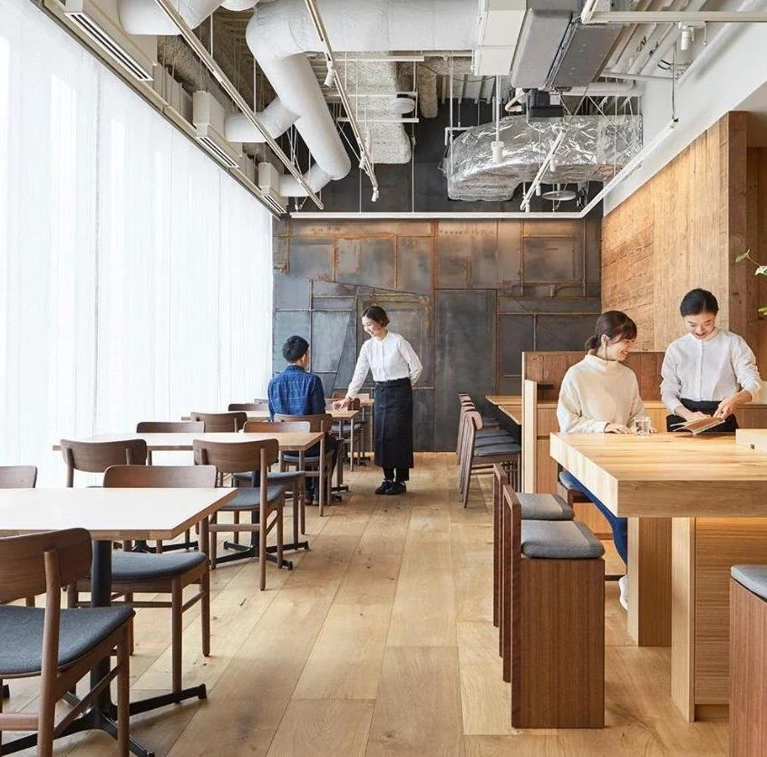 A modern, minimalist cafe with light wooden floors and furniture, concrete and exposed ceiling pipes, and large white curtains covering windows. There are four staff members and no customers visible.