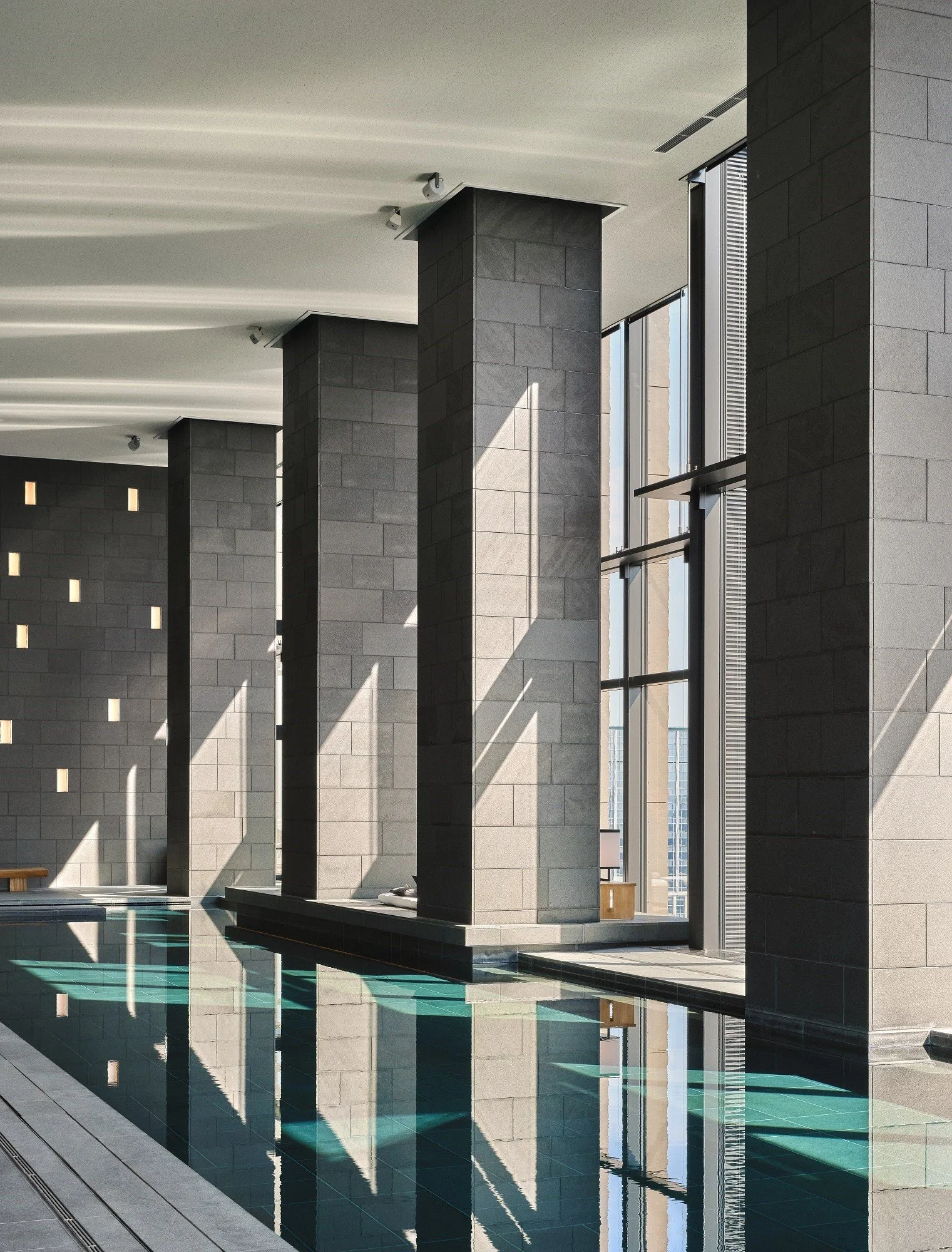 Interior of a modern indoor swimming pool with large stone columns, high windows, and reflections in the water.