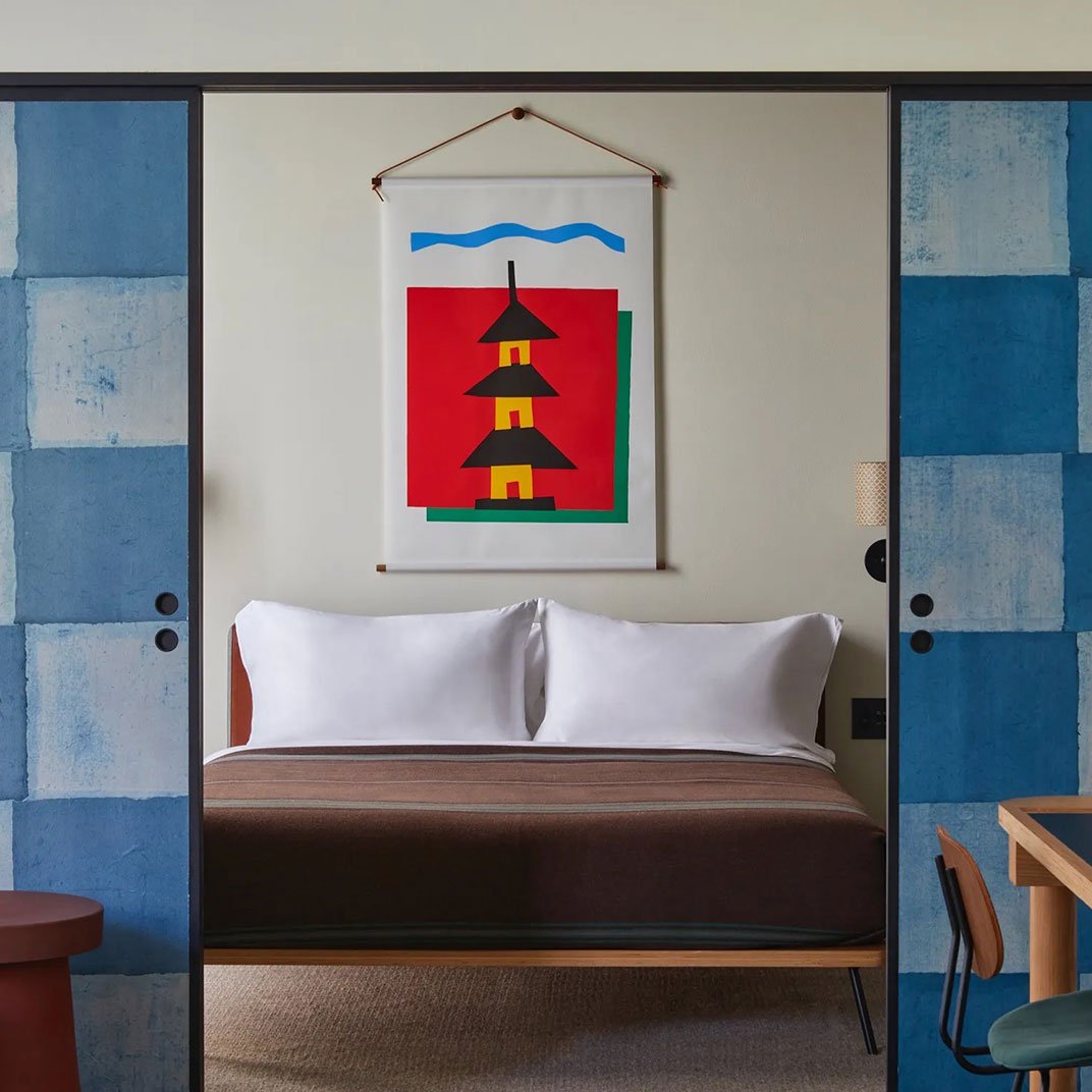 A hotel room with a bed, white pillows, and a colorful modern painting of a pagoda and water above the bed, framed by blue sliding doors.