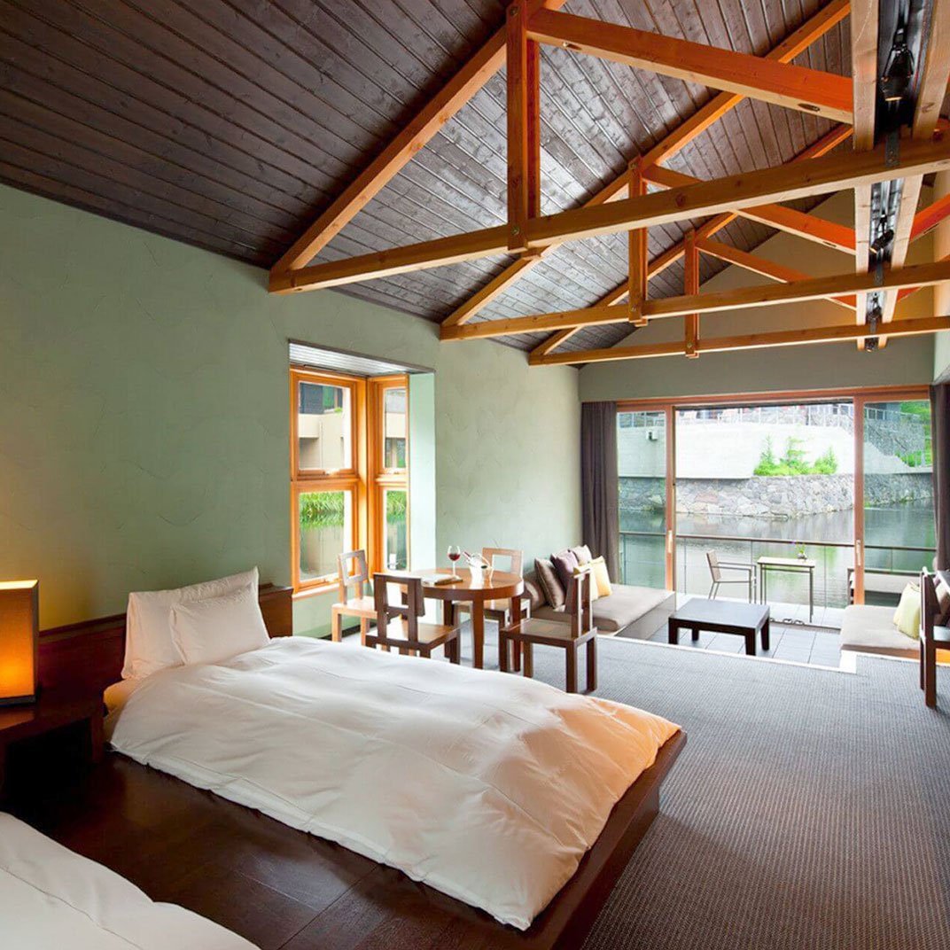 A hotel room with two beds, a dining area, and a seating area with a view of a balcony overlooking water and greenery.