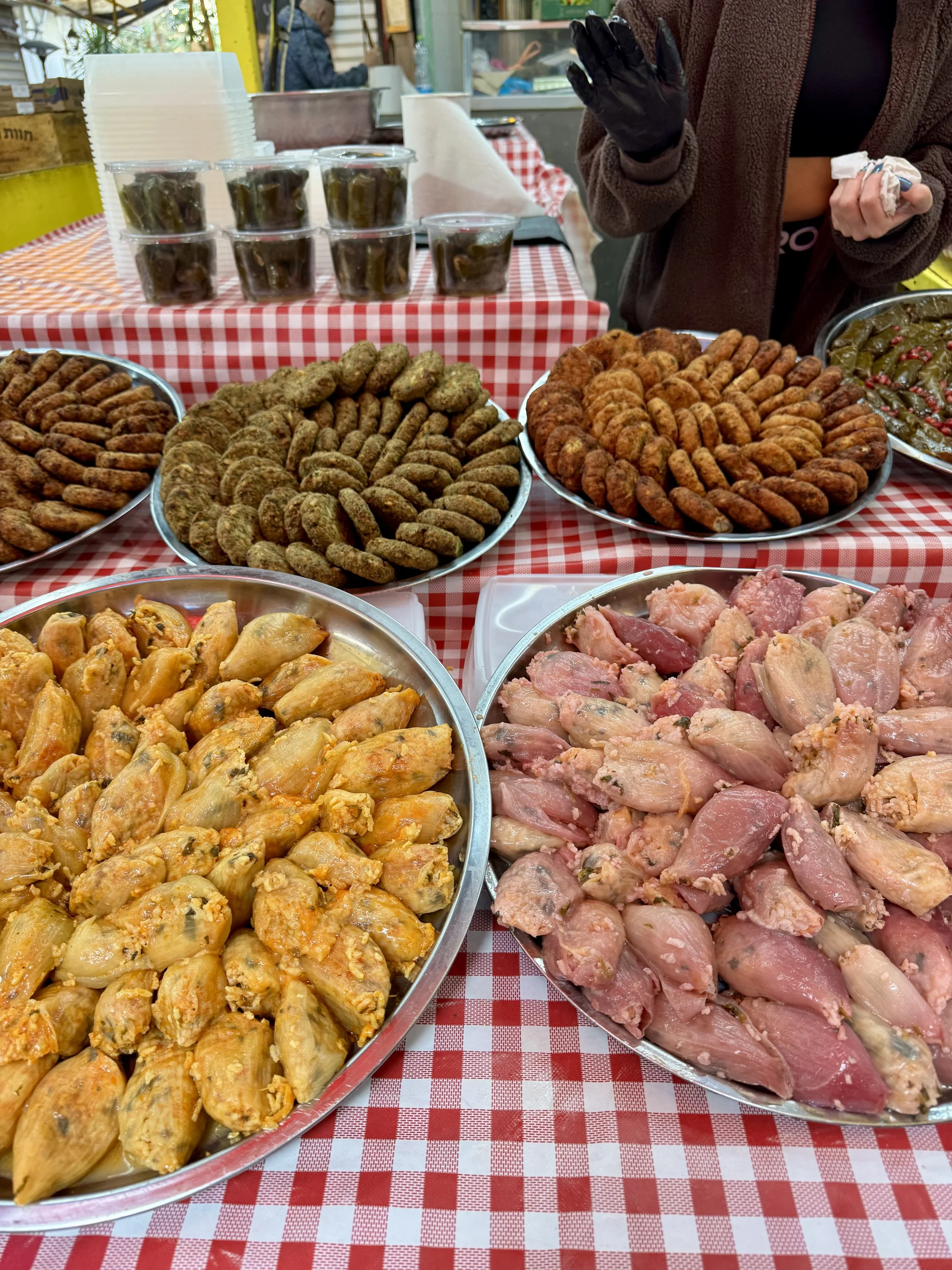 A variety of plates with cooked meats, pickled vegetables, and containers of peppers displayed on a table with a red and white checkered tablecloth at a food market.