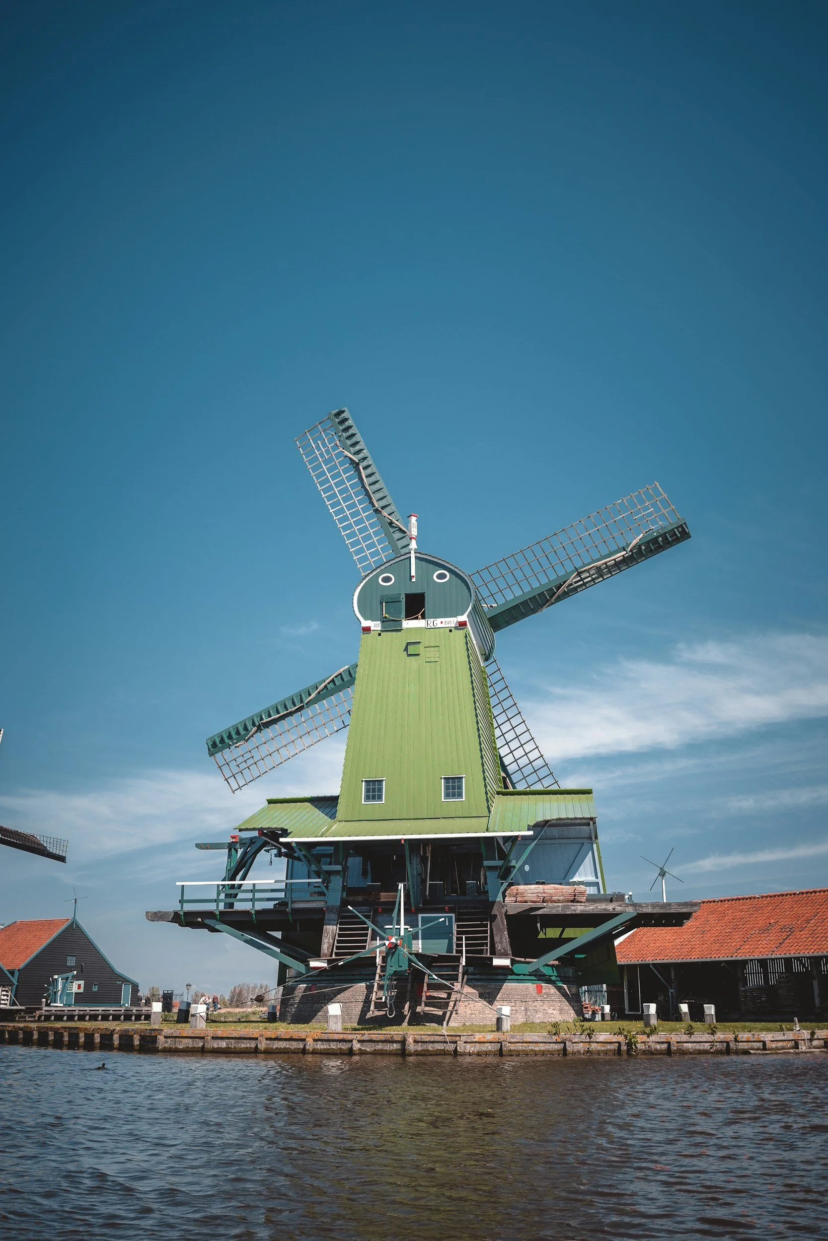 Green traditional Dutch windmill beside water under a blue sky with some clouds.
