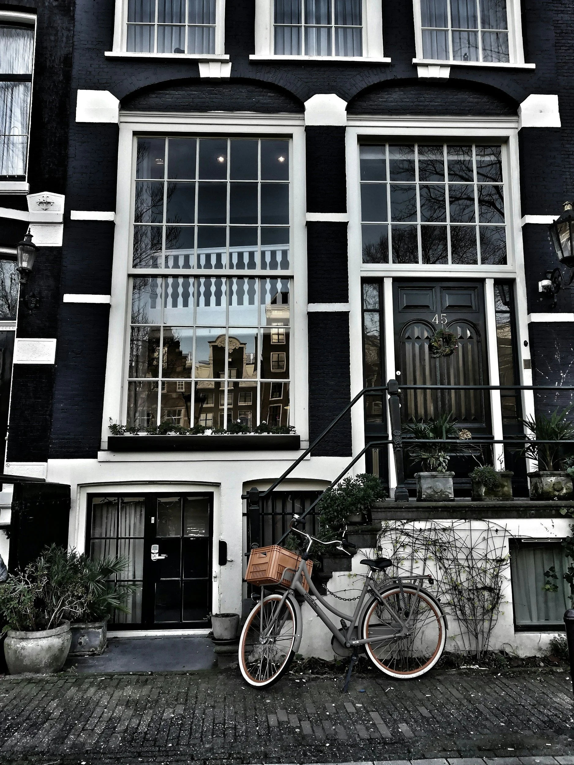 A black and white multi-story building with large windows, a black front door with the number 45, and a bicycle leaning against the stairs. Potted plants are on the stairs and outside the basement door.