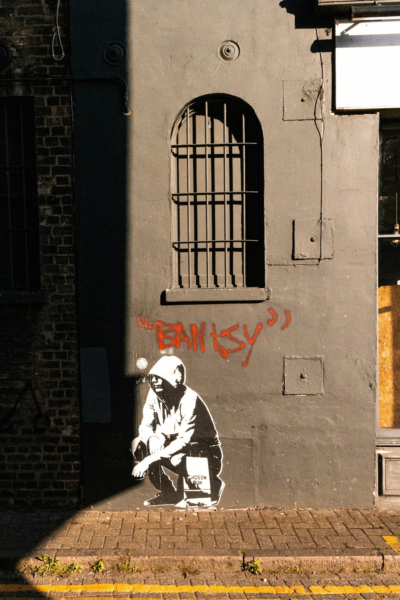 Street art graffiti depicting a person squatting, wearing a hoodie and mask, with the words 'BANSY' written above in red spray paint.