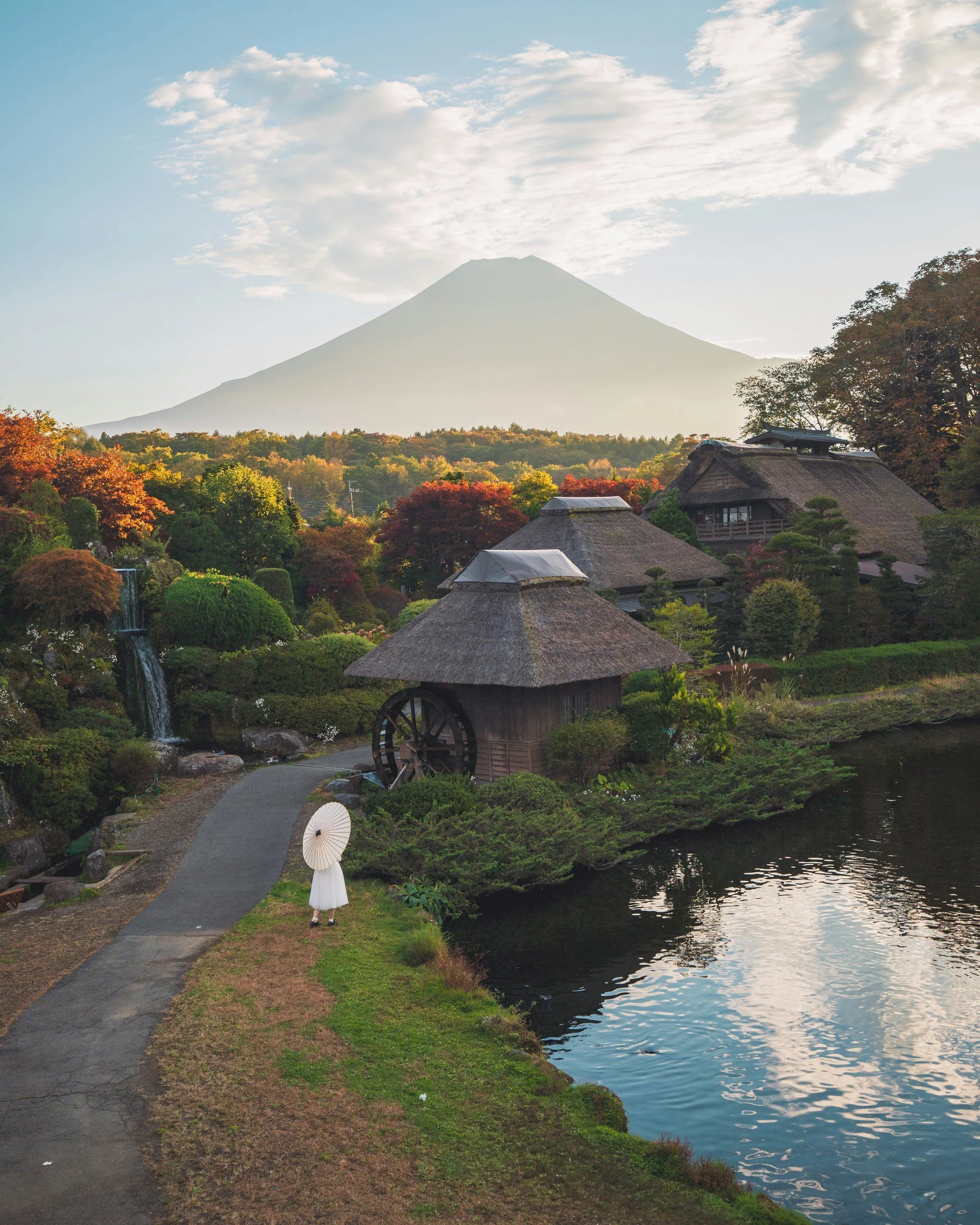 A serene landscape with traditional Japanese thatched-roof buildings, a small waterfall, lush trees with autumn foliage, a pond reflecting the sky, and Mount Fuji in the background under a partly cloudy sky.