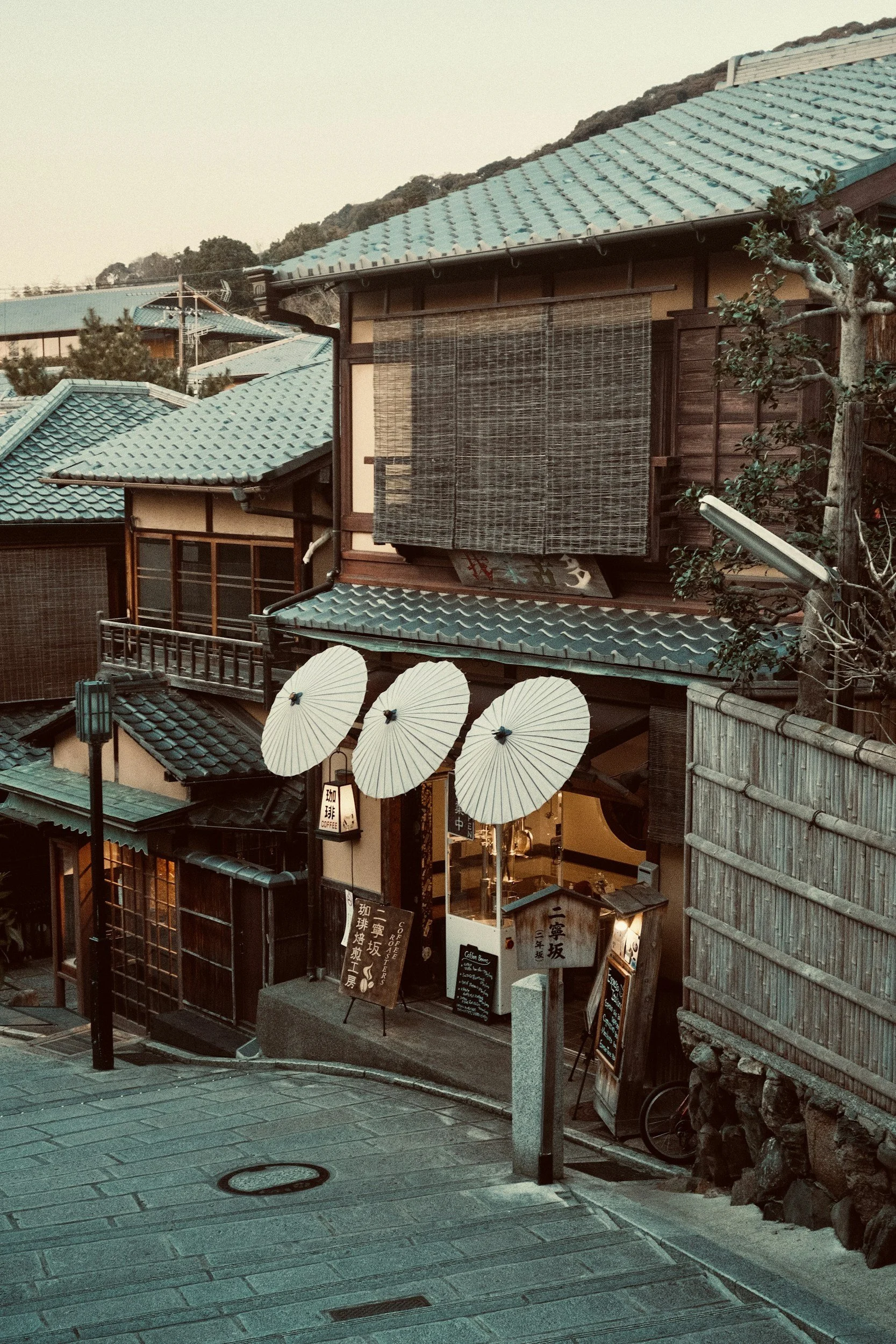 Traditional Japanese building with teal tiled roof, paper umbrellas at entrance, and hanging bamboo blinds, on a quiet street at dusk.