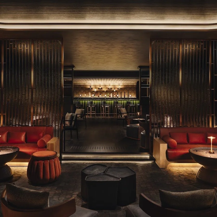 A dimly lit upscale bar with a seating area in the foreground featuring red sofas and tables, and a bar with stools in the background illuminated by warm lighting.