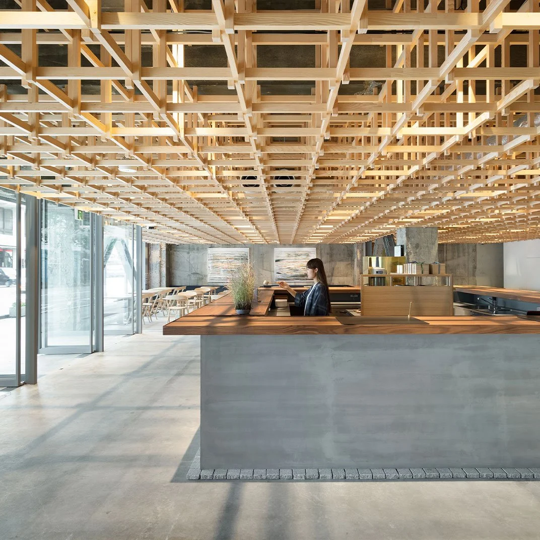 Modern cafe interior with wooden ceiling grid, concrete walls, and a wooden counter where a woman is sitting and working on a laptop. Large glass doors allow natural light to fill the space.