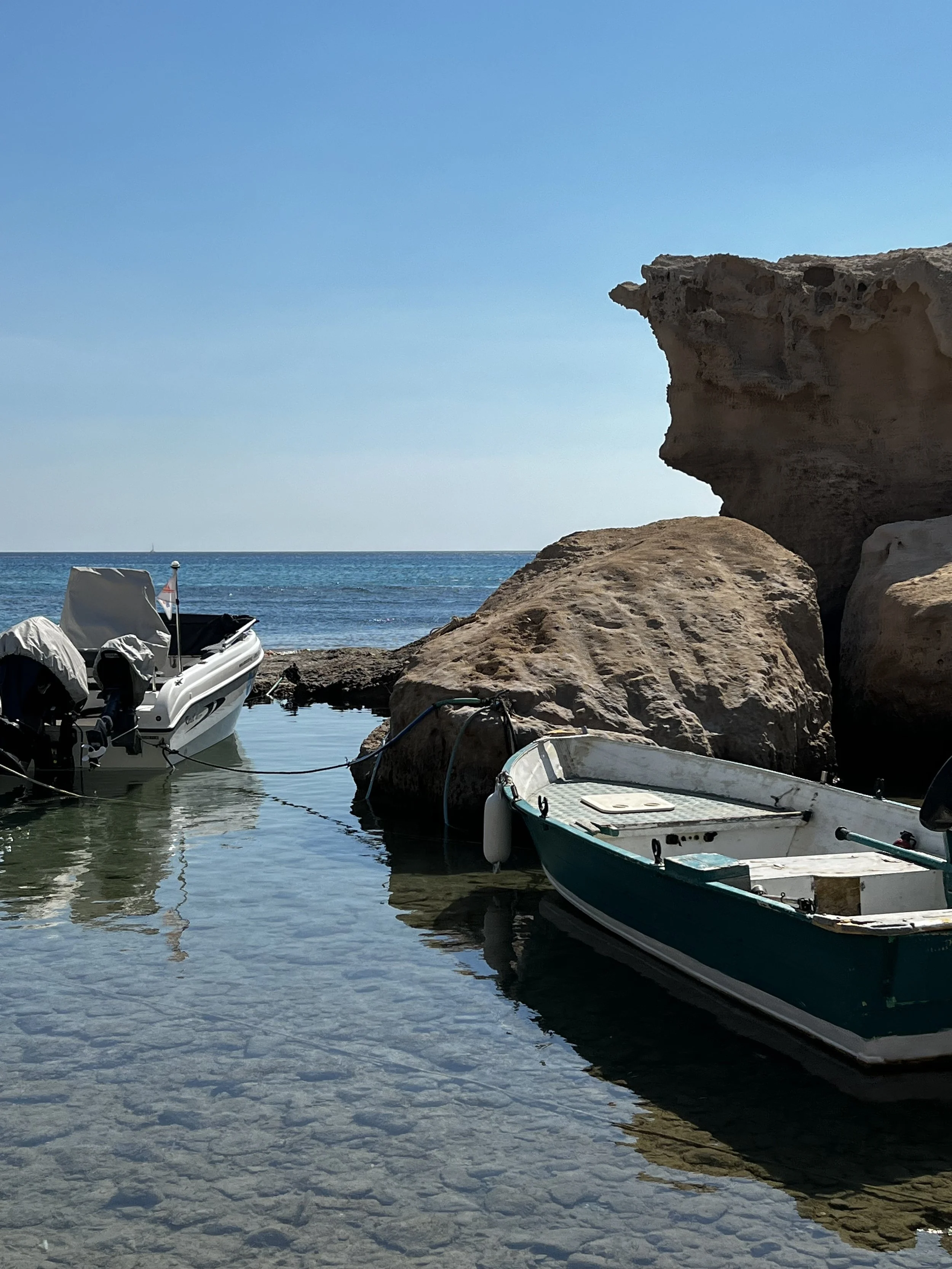 Small boats moored near large rocks on a calm sea, with a clear blue sky overhead.