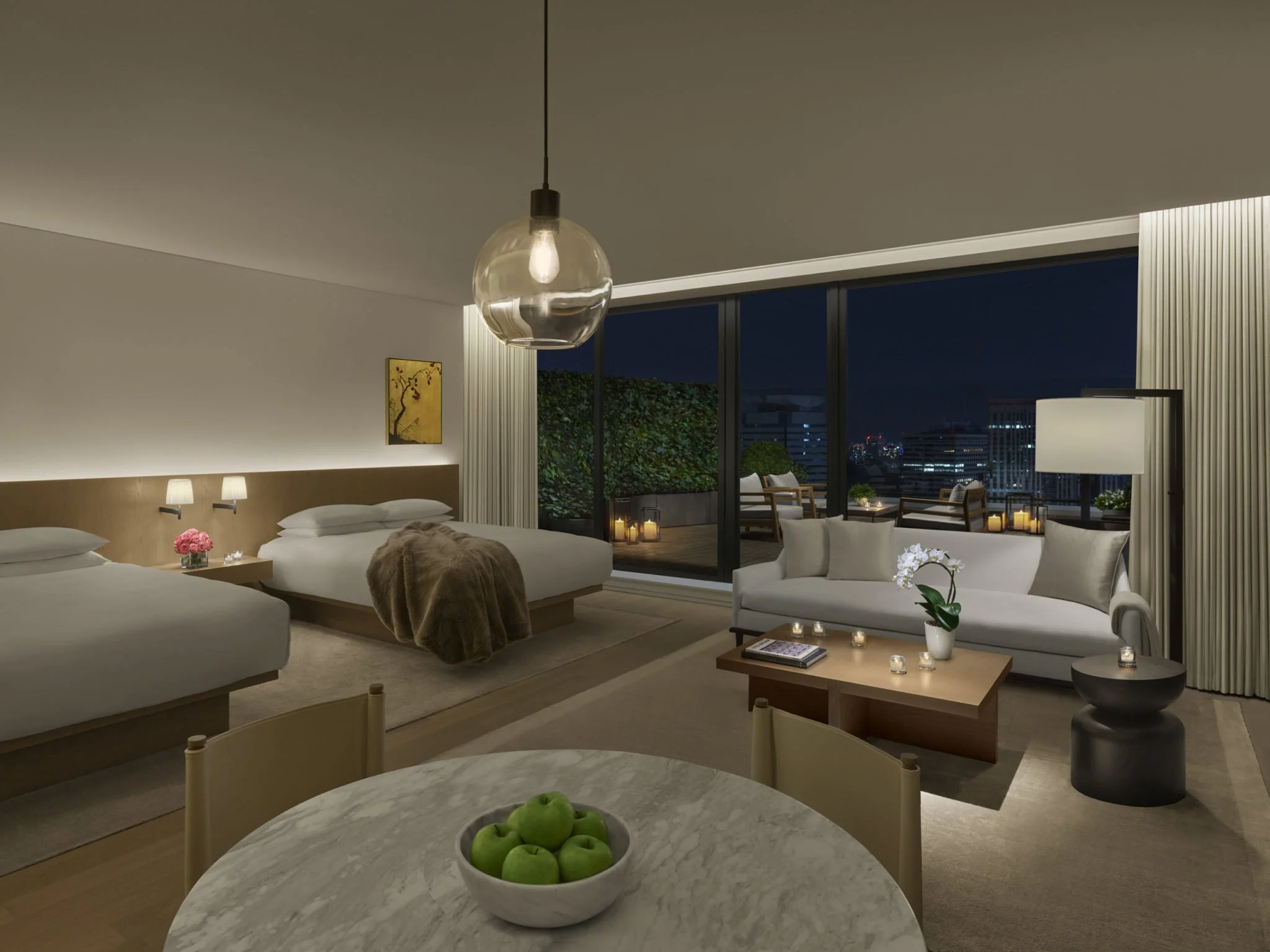 Modern hotel room with two beds, a sitting area with a sofa, and a large window showing a city skyline at night. Decor includes candles, flowers, and a bowl of green apples.