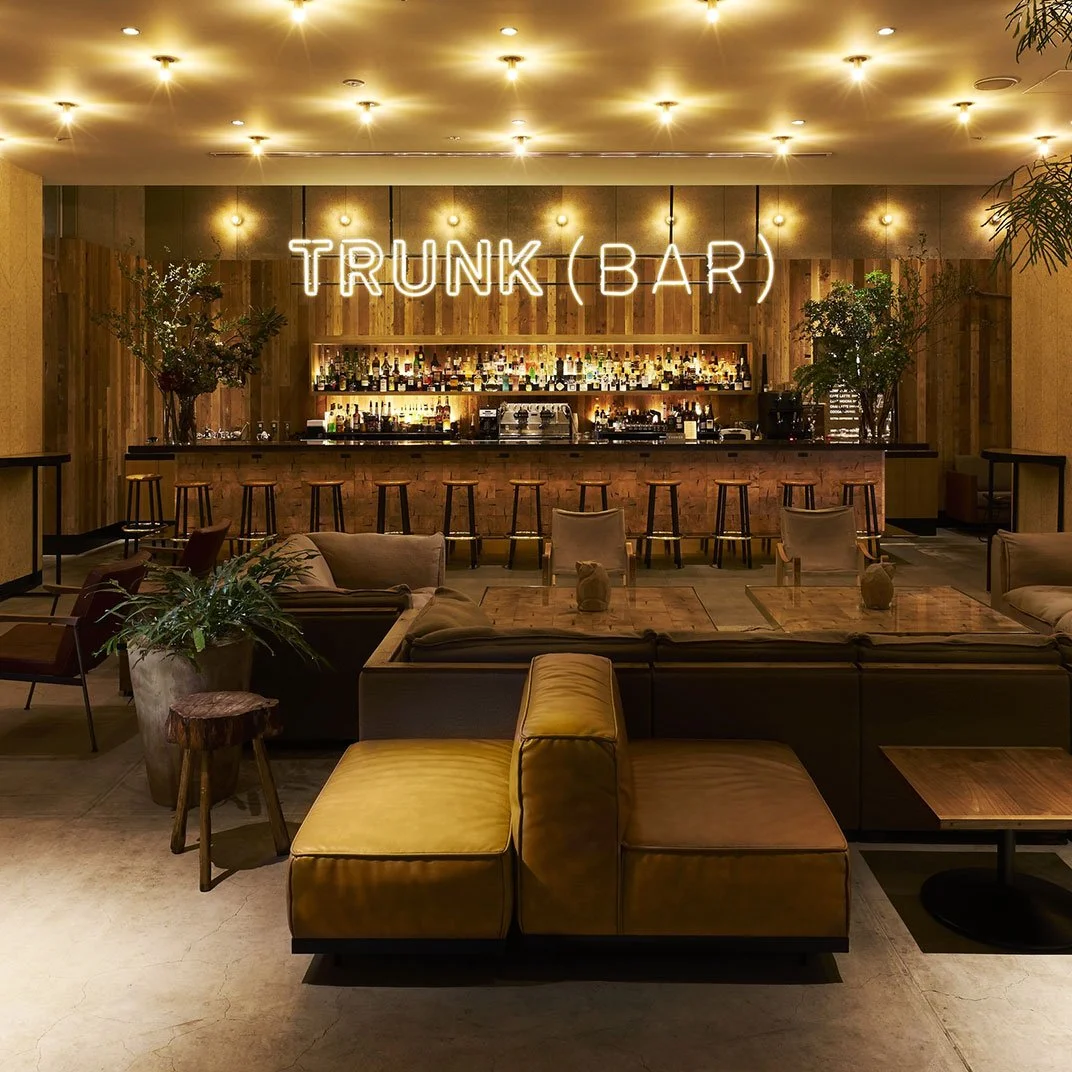 Interior of a bar named 'TRUNK BAR' with a neon sign, various bottles of alcohol on shelves, seating, and decorative plants.