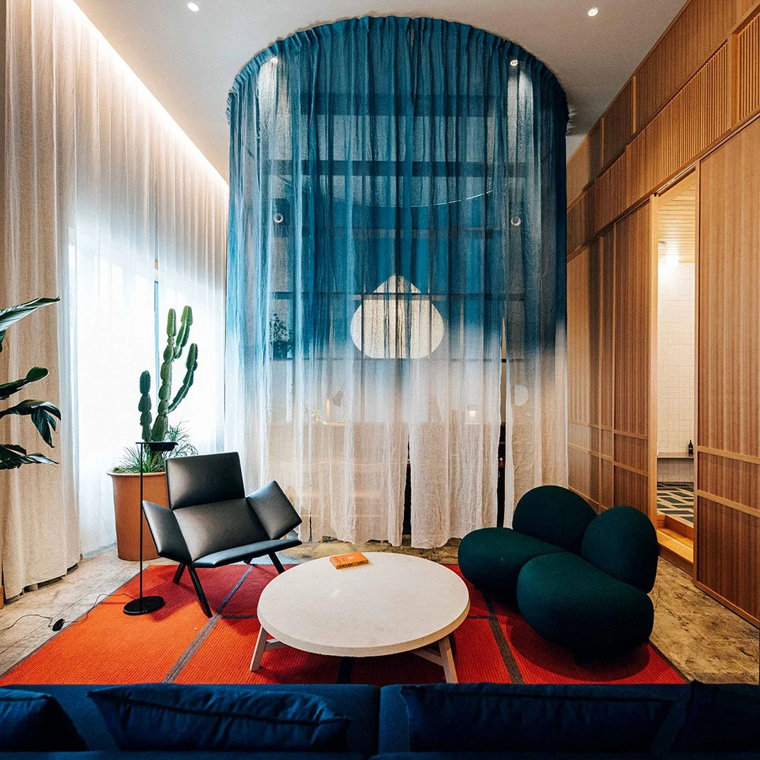 Contemporary living room with black leather chair, dark green curved sofa, white round coffee table with an orange book, potted cactus plant, orange and red area rug, and wooden wall panels, separated by sheer blue curtains.