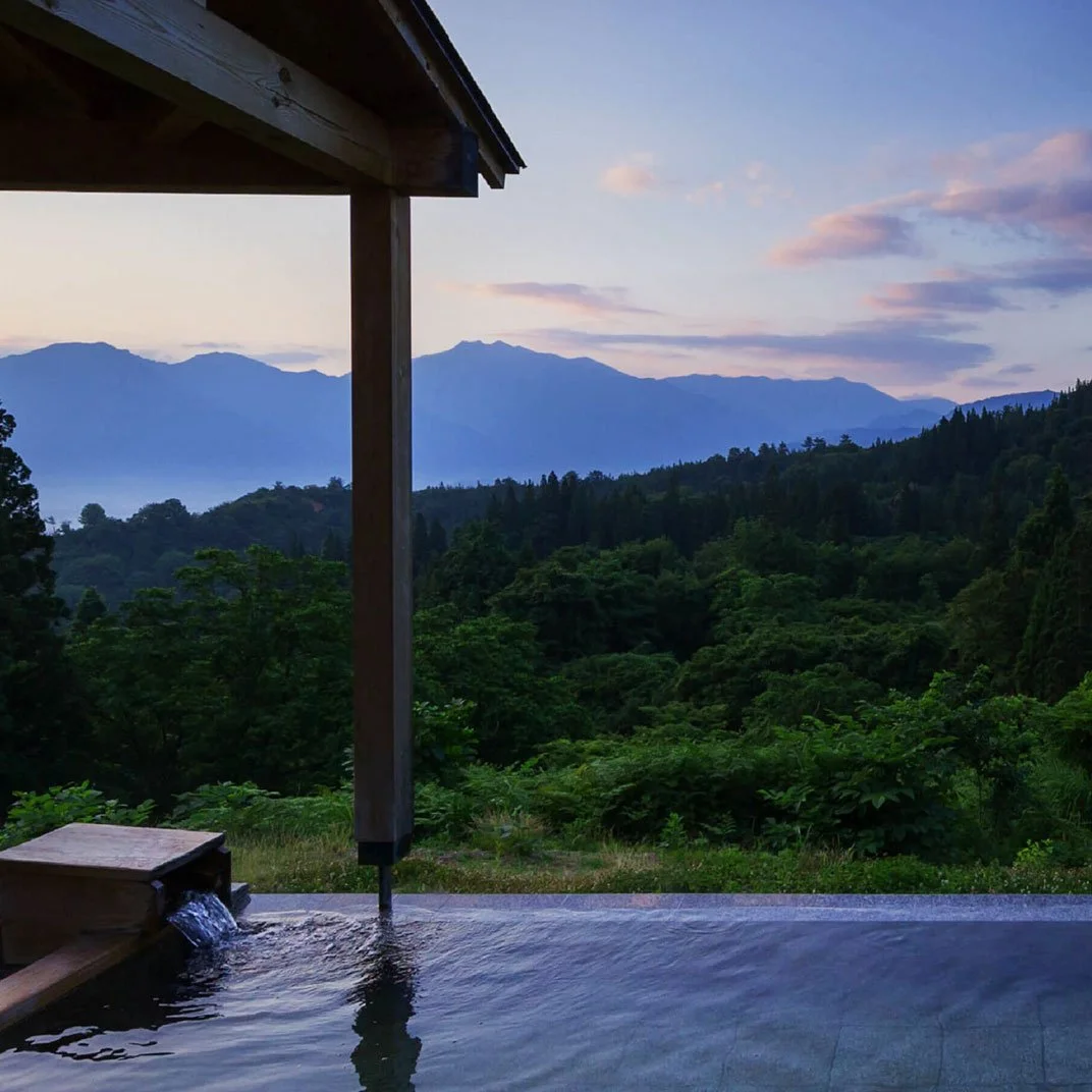 A scenic view of a mountain range with green forests and a cloudy sky, viewed from an outdoor hot spring bath with wooden structure and flowing water.