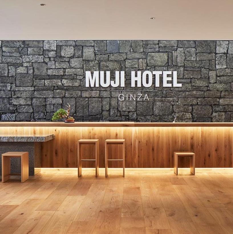 Lobby of MUJI Hotel Ginza with a stone wall behind a wooden counter and three stools, along with a small table and plant.