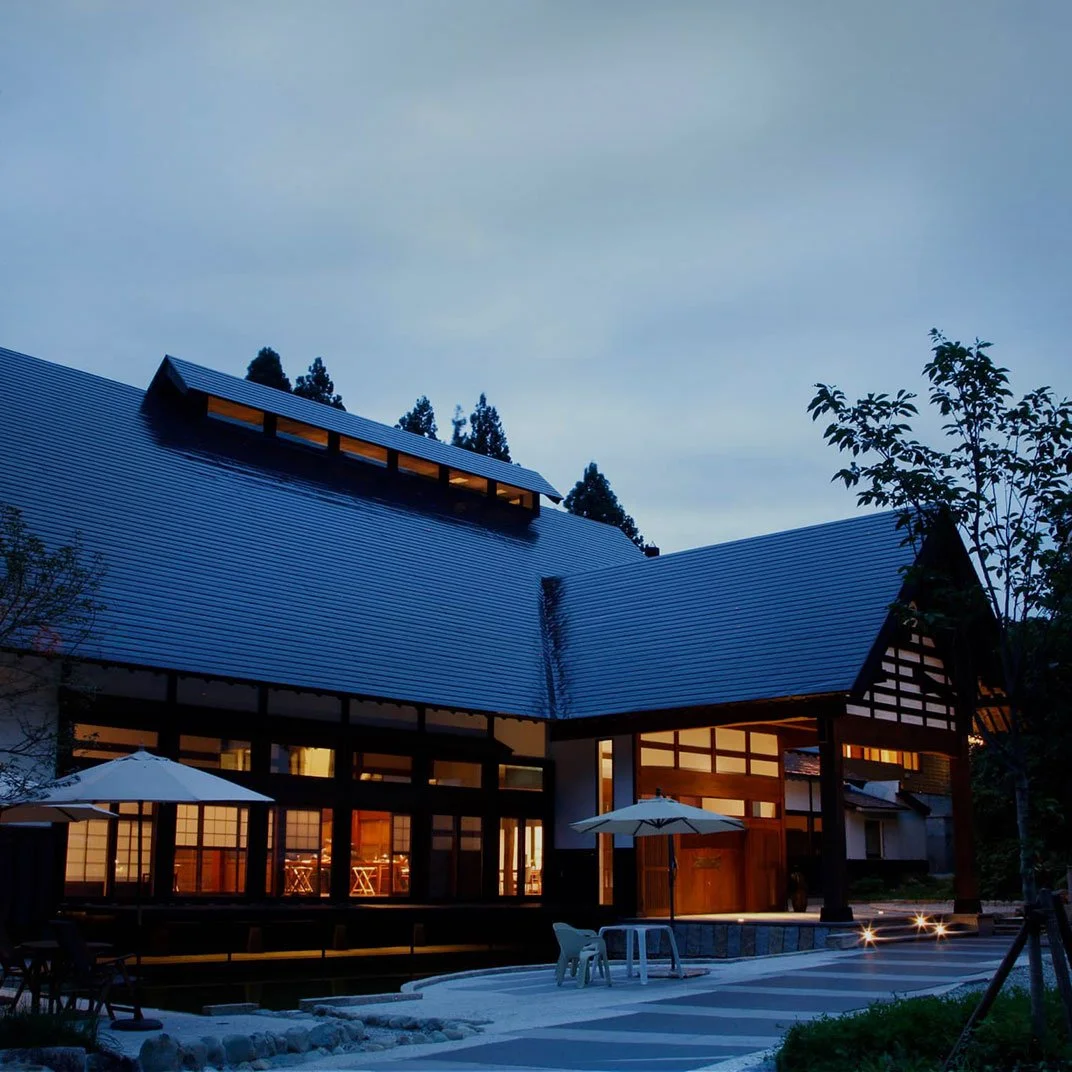 A house with a traditional Japanese architectural style, featuring dark wooden framing, large windows, and a sloped roof, illuminated from inside during dusk or early evening.