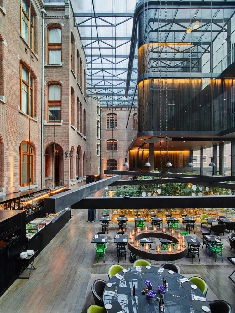 Indoor dining area at a modern restaurant with a glass ceiling, brick walls, and stylish black and green chairs arranged around tables, including a large round table with floral decorations.