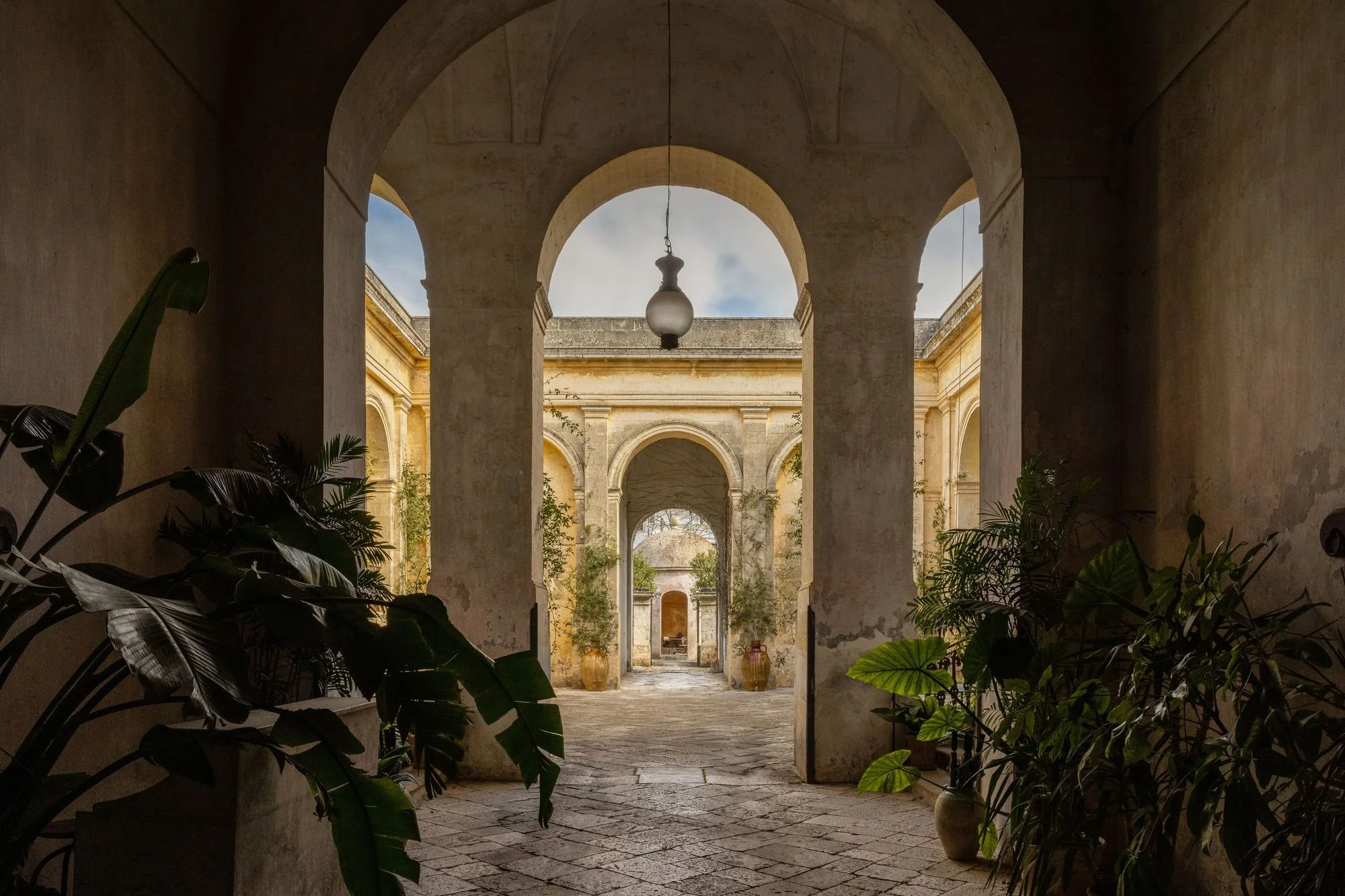 View through arched doorways of a historic building with stone walls, plants, and a sky with clouds visible in the background in Italy.